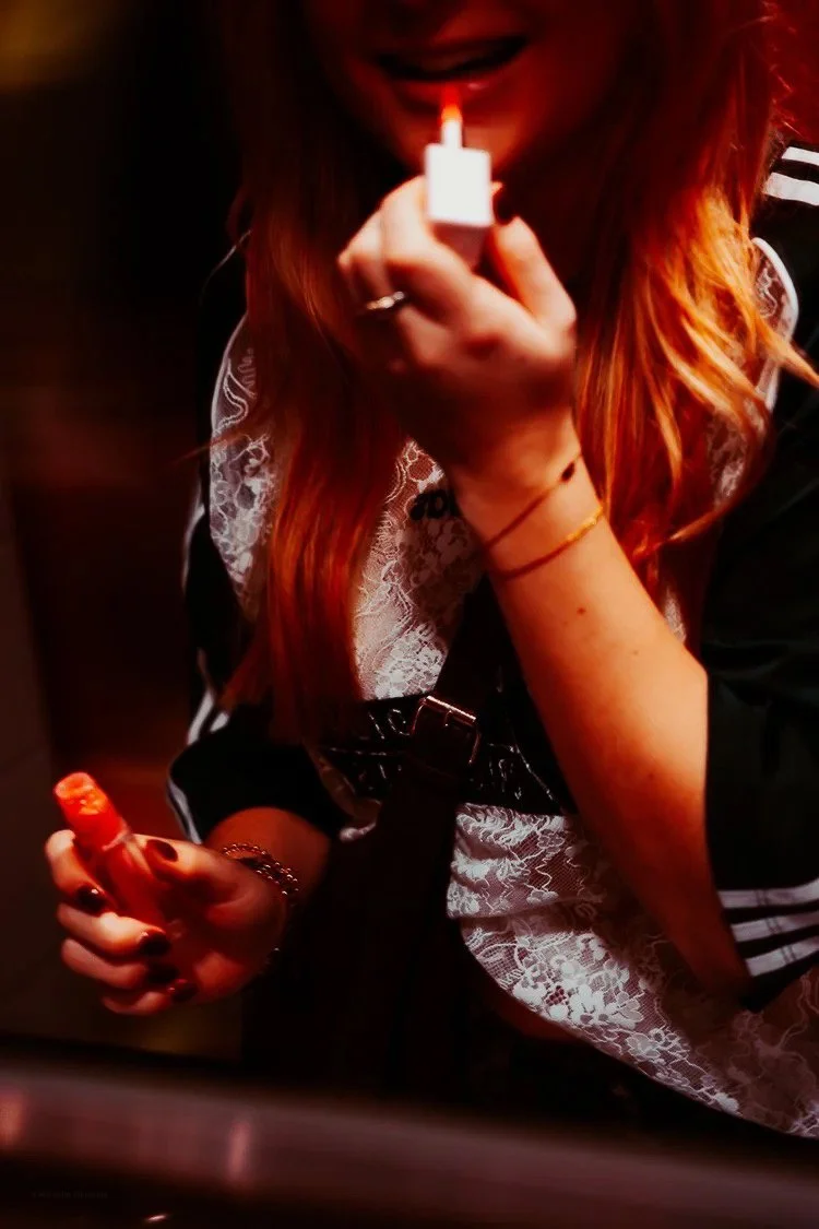 A woman with long red hair holding a lighter and a cigarette lighter in a dimly lit environment. She is smiling slightly, with her face partially visible, and wearing a black and white lace and striped shirt.