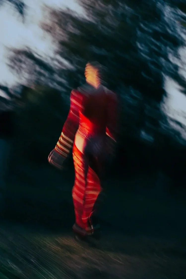 Blurry image of a woman walking on a road at sunset, wearing a jacket and striped pants, with trees in the background.