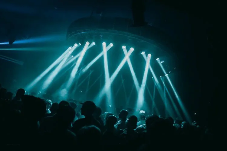 Crowd inside a dark nightclub with bright white stage lights overhead.