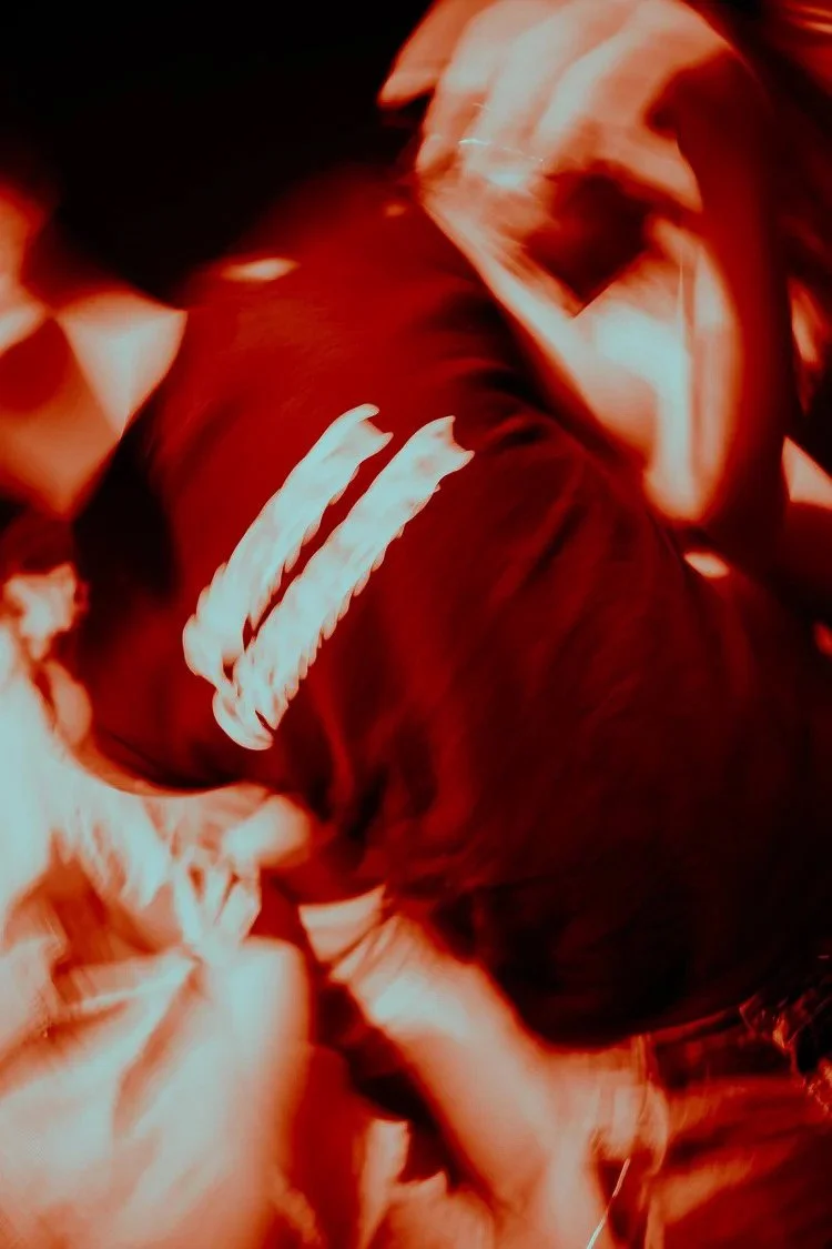 A person with long hair wearing a jacket with white stripes on the sleeve, sitting or leaning in a dimly lit environment with red lighting.