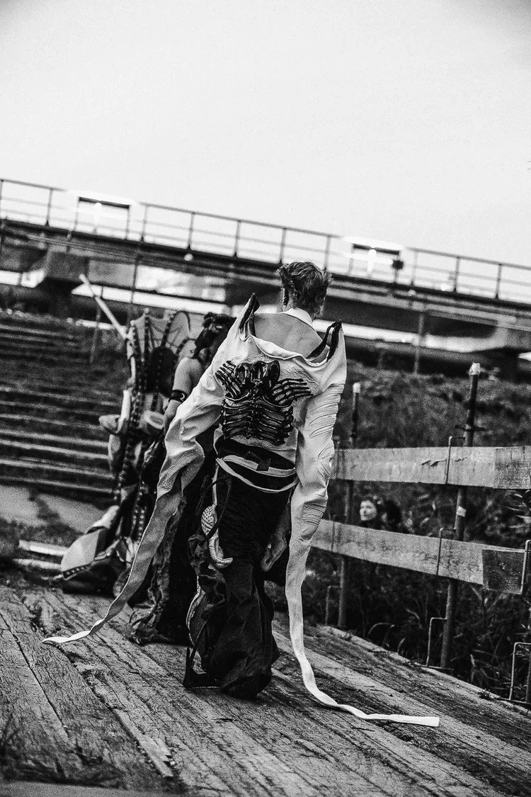 A person walking down a wooden pathway carrying a costume with a skeleton design on the back, with other people and a staircase in the background.