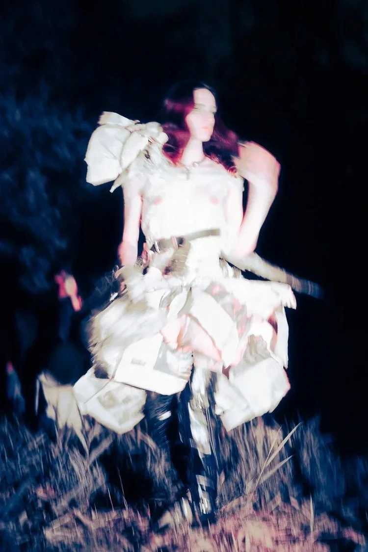 A person is standing outdoors at night, wearing an elaborate outfit made of layered, patchwork-style fabric. The outfit has large, ruffled details, and the person has long dark hair with reddish highlights.