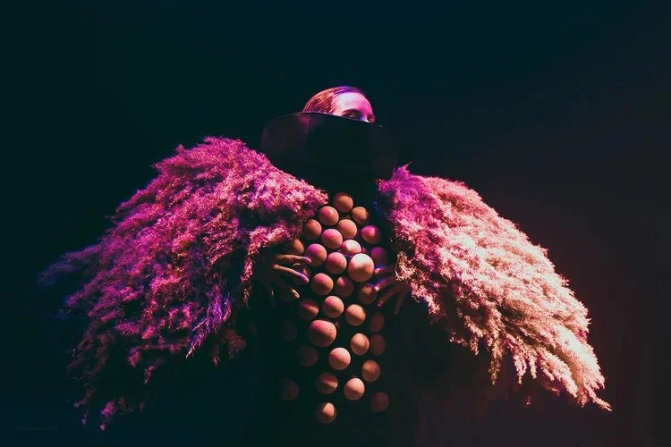 A person wearing an avant-garde costume with large pink and beige fluffy wings, oversized beige beads, and a black face mask or visor, standing against a dark background.