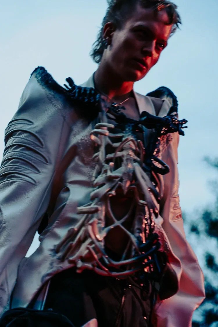A person with short, styled hair wearing a light-colored, shiny jacket with dark accents and black laces, standing outdoors during dusk or dawn.