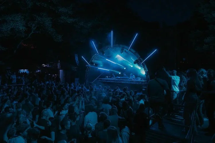 At a nighttime outdoor concert, a large crowd watches a DJ perform on a stage lit with blue and white lighting.