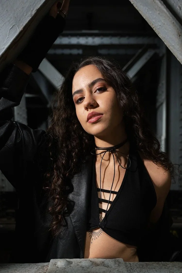 A woman with long curly hair, wearing a black top with criss-cross straps and a black choker, posing under a metal structure.