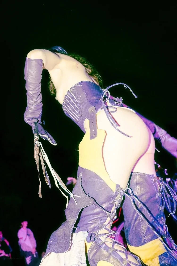 Person wearing black leather outfit with purple gloves and yellow accents, striking a stylish pose against a dark background.