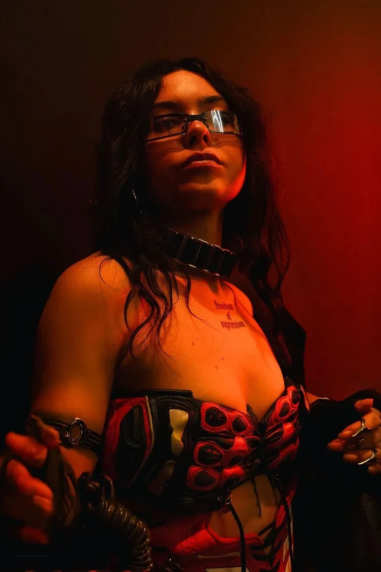 A woman with black, curly hair, glasses, and multiple accessories wears a black and red outfit and stands in a dimly lit red-hued room.