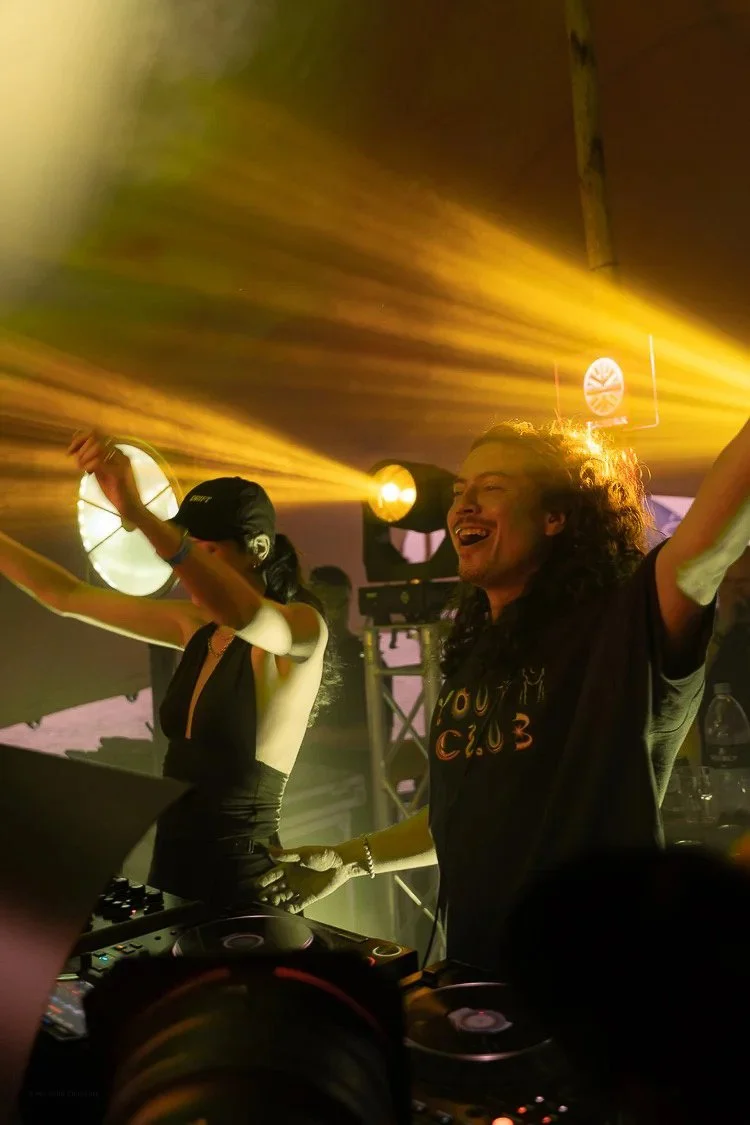 Two DJs performing at a nightclub, with yellow and orange stage lighting and a happy, energetic atmosphere.