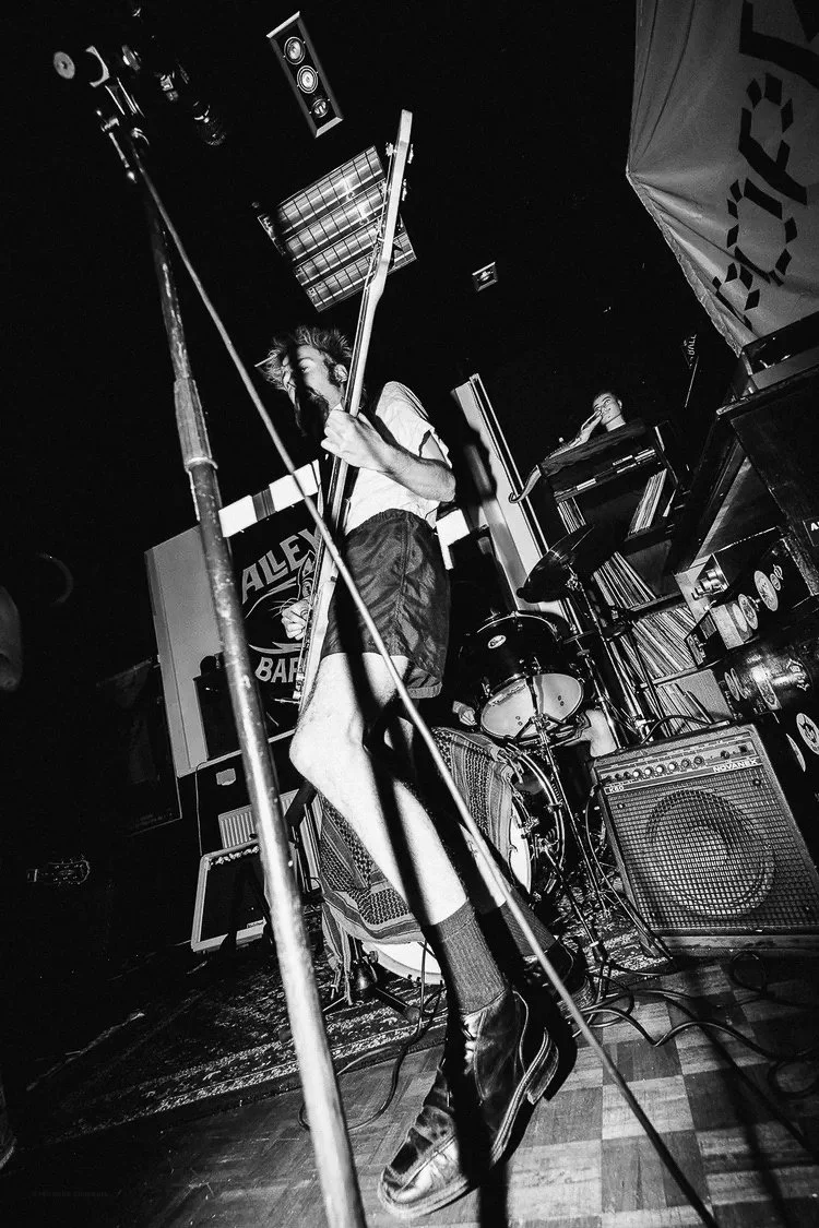 A black and white photo of a live music performance with a male singer holding a microphone stand, wearing shorts, a T-shirt, and boots, standing on a stage with musical equipment around him. A keyboard player is visible in the background. The settin