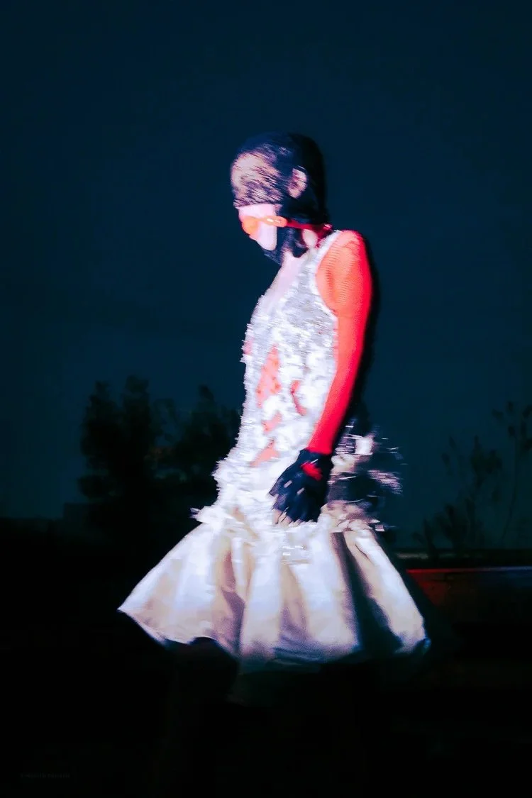 Person in a fancy gown and gloves walking outdoors at night, wearing a face mask and head covering.