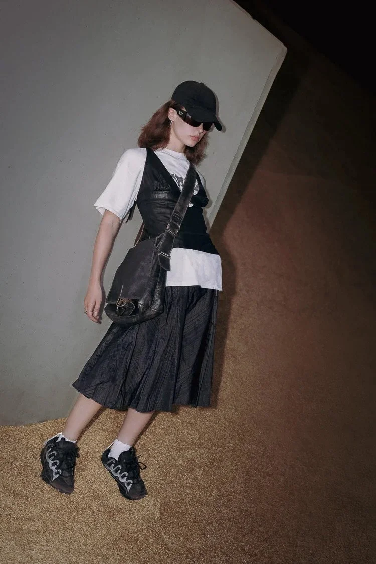 A woman wearing a black cap, black sunglasses, a white t-shirt, a black vest, a black pleated skirt, and black sneakers with white socks, carrying a black shoulder bag, leaning against a wall.