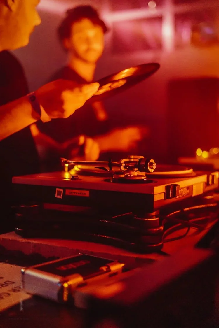 A DJ plays music on turntables in a dimly lit setting, with a smiling man in the background.