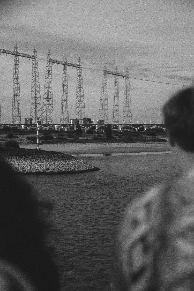 Black and white photo of power lines and transmission towers near a body of water with people in the foreground.