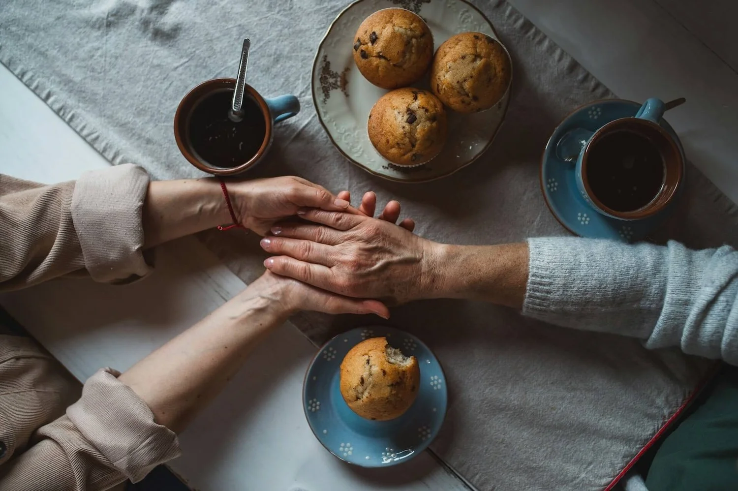 Two people holding hands across a table with tea and muffins, reflecting compassionate support, care and meaningful conversations.