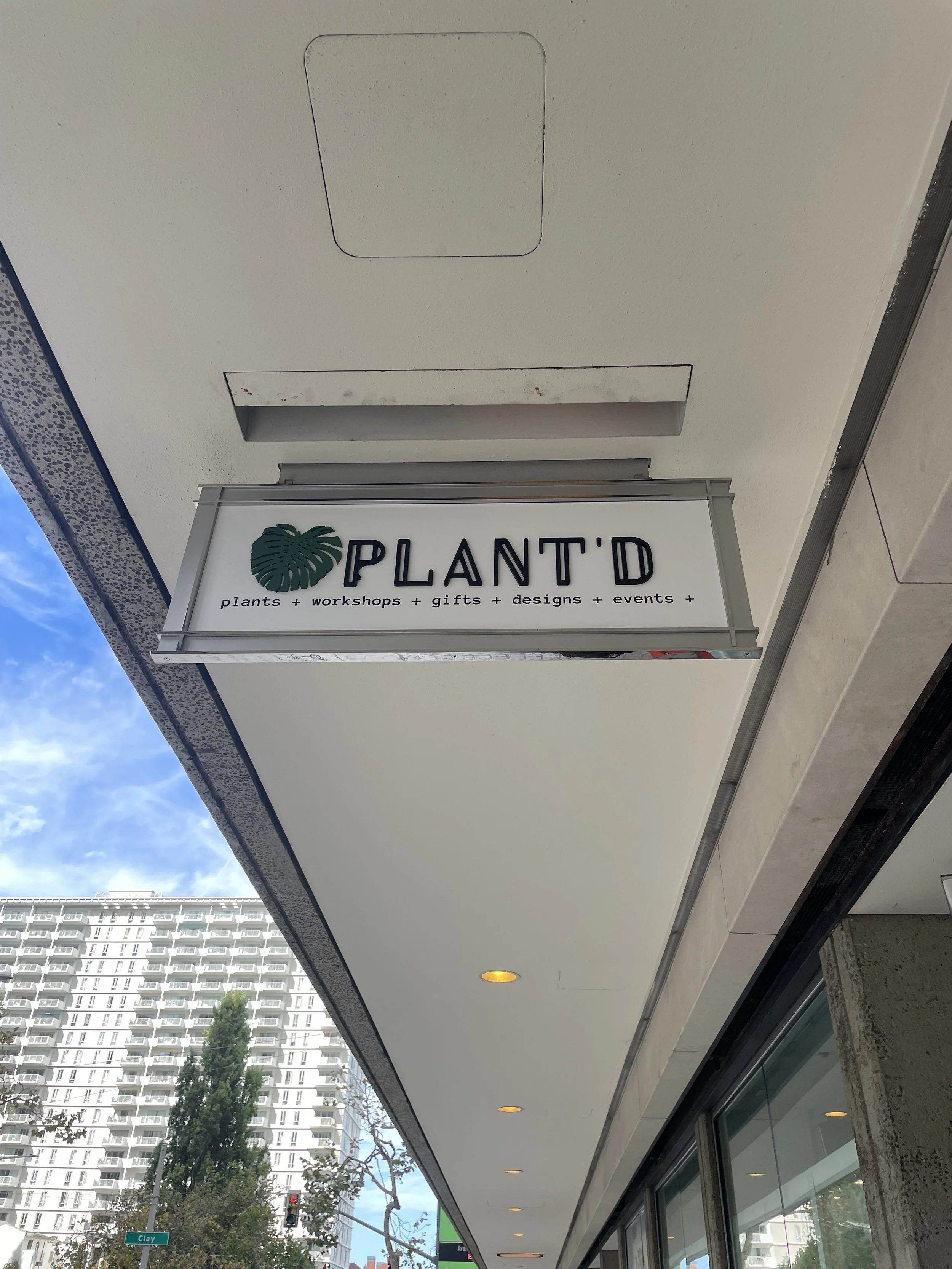A hanging sign for a business called PLANT'D, offering plants, workshops, gifts, designs, and events, attached to the exterior of a building in an urban area with a high-rise in the background.