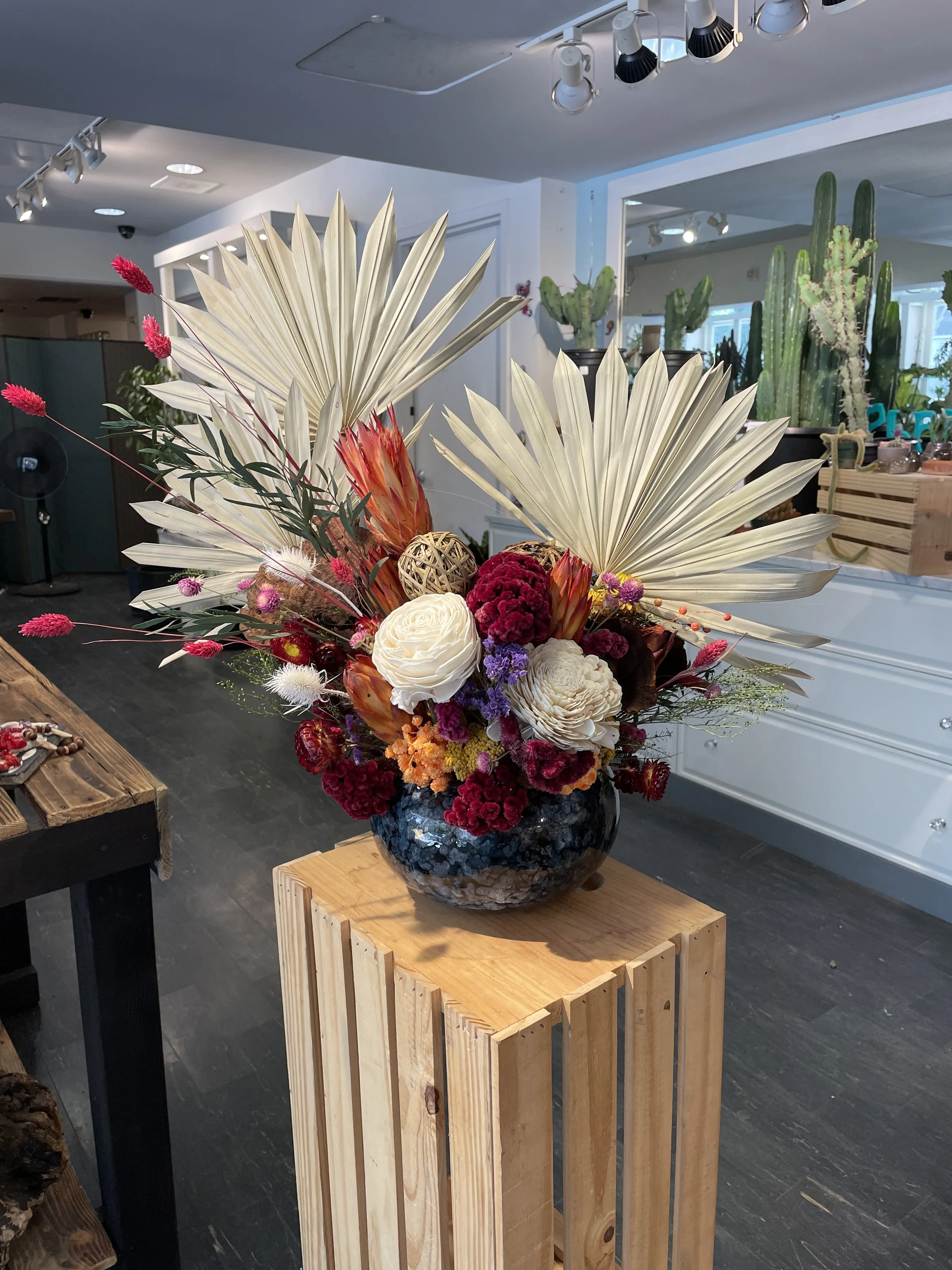 A flower arrangement in a black textured vase placed on a wooden stand. The arrangement includes dried leaves, bright pink flowers, white roses, red and orange flowers, purple, and other colorful blooms with cactus plants in the background.