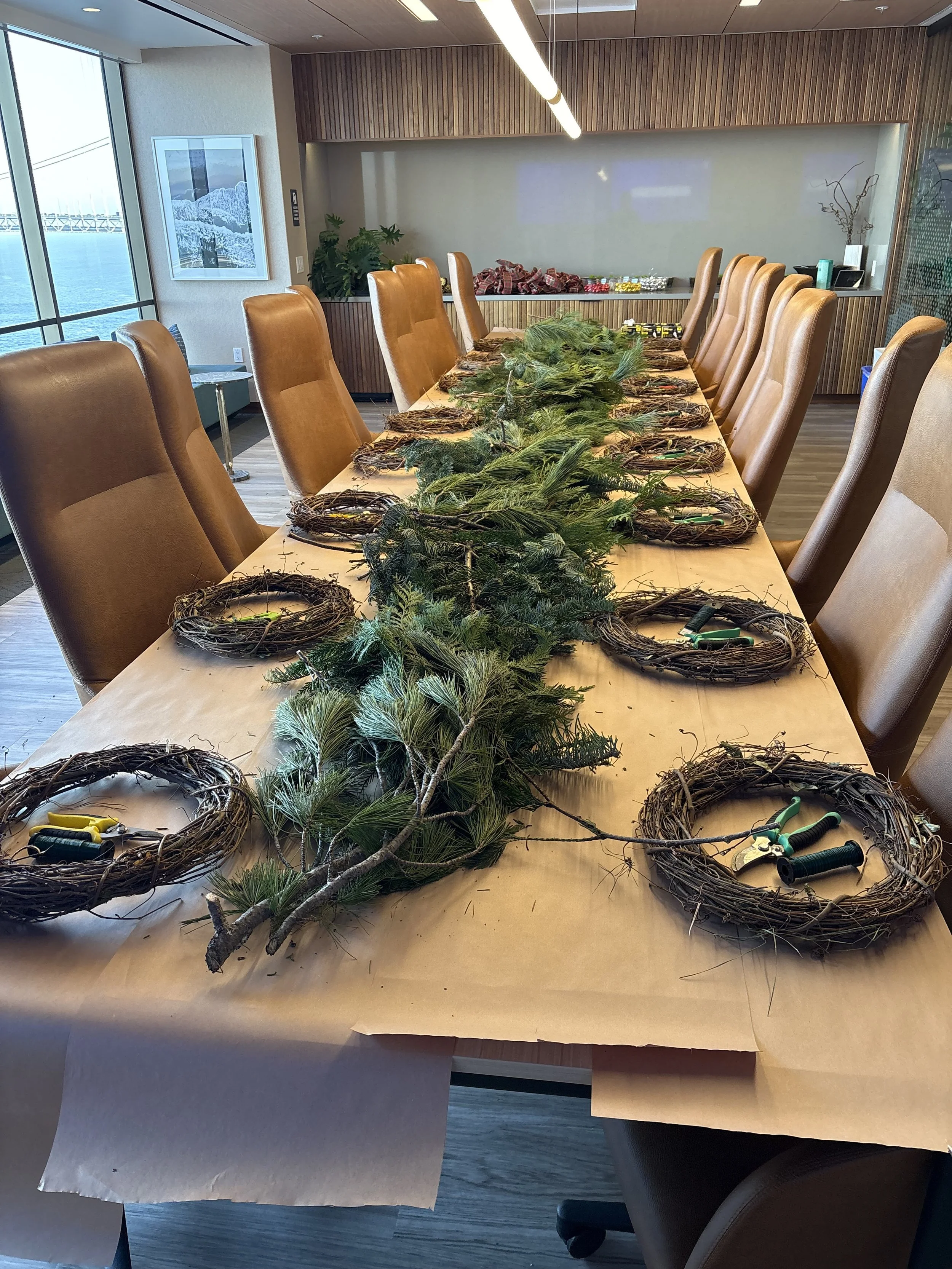 A table with wire wreath bases, pine branches, and floral tools, ready for Christmas decoration, in a conference room with brown chairs and a window view of a bridge over water.