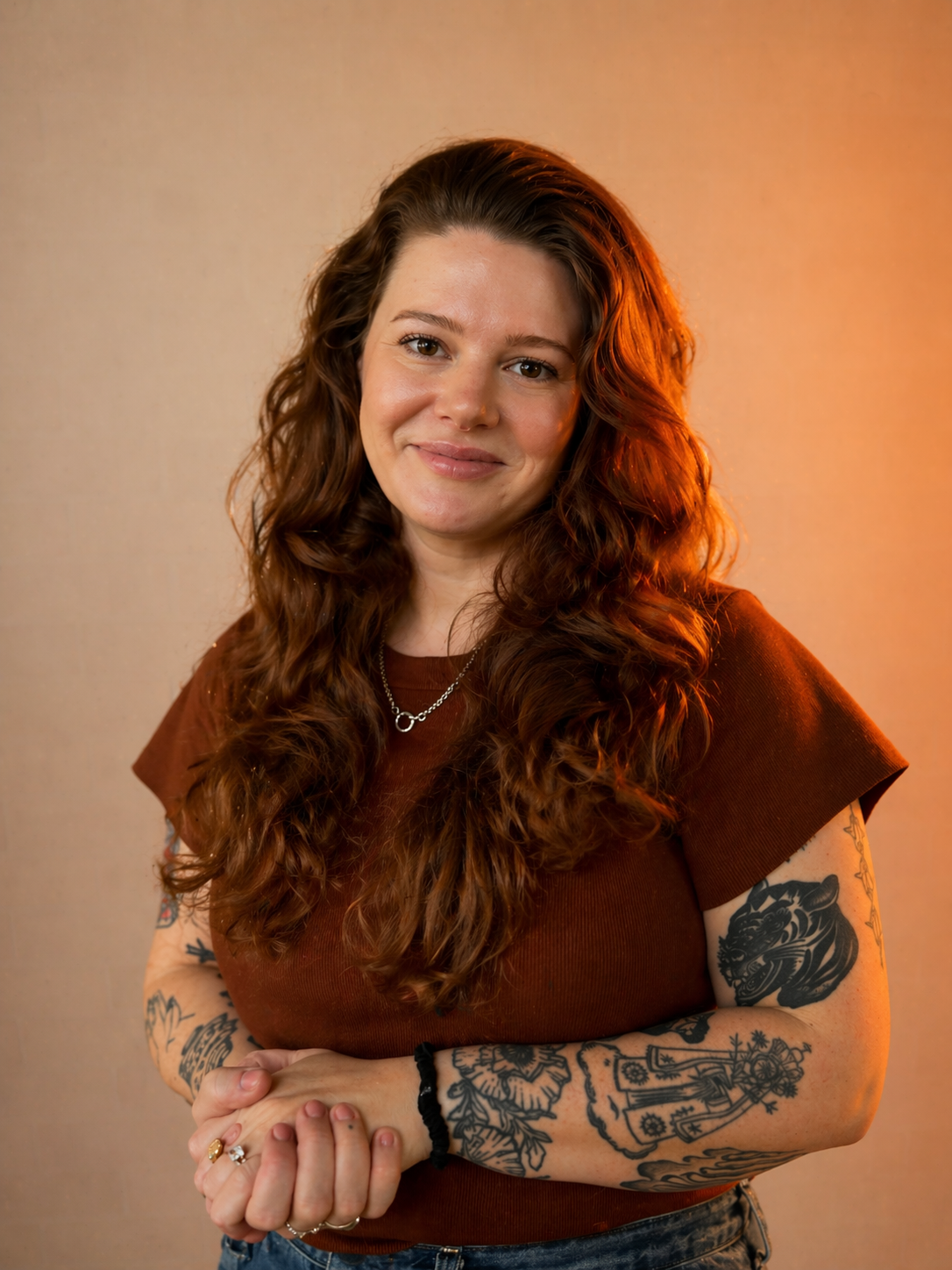 Portrait of a woman with long, curly red hair, wearing a brown T-shirt, silver necklace, and rings, standing against a neutral background with warm lighting.