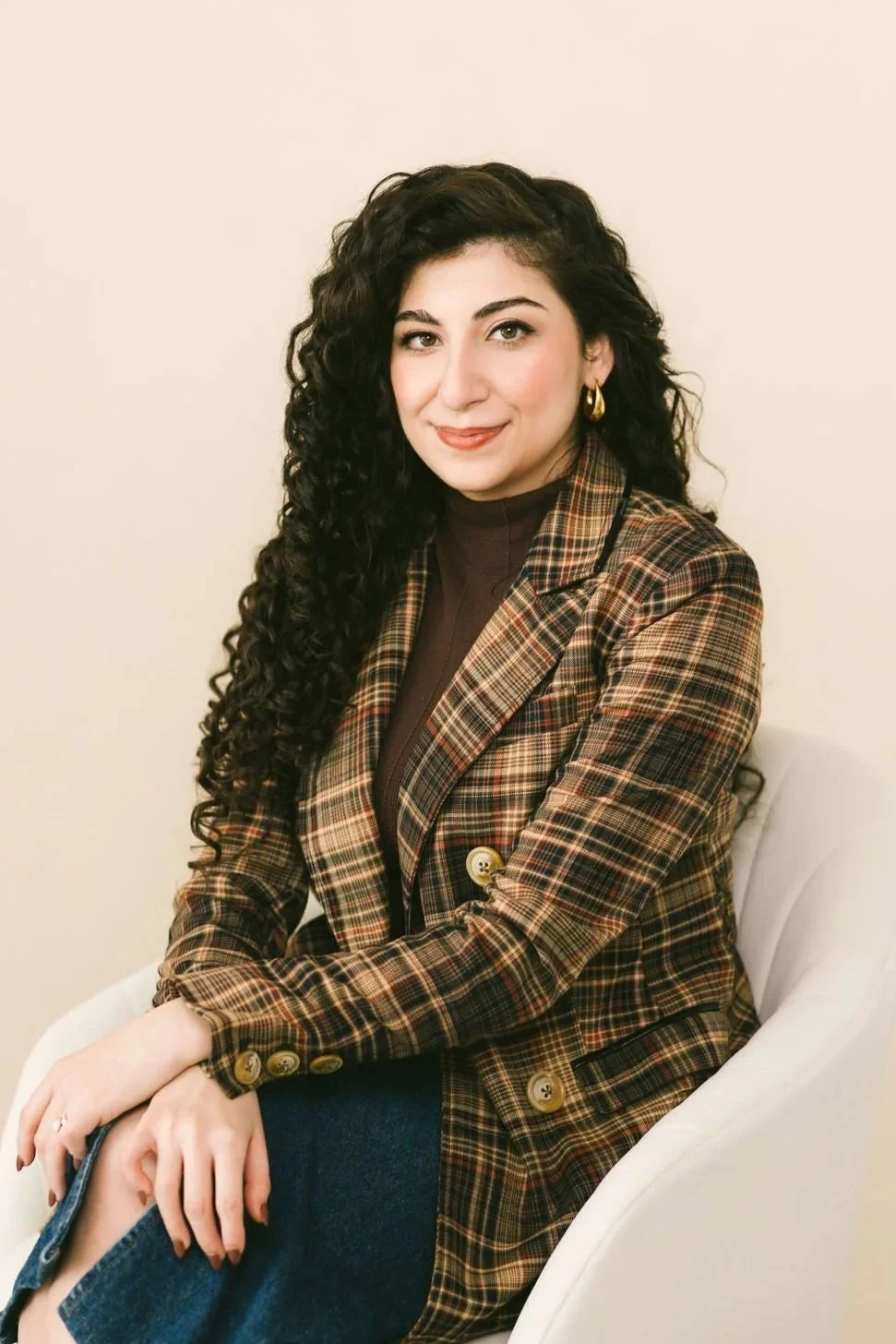 A woman with long, curly dark hair and light skin, smiling while sitting in a beige chair.