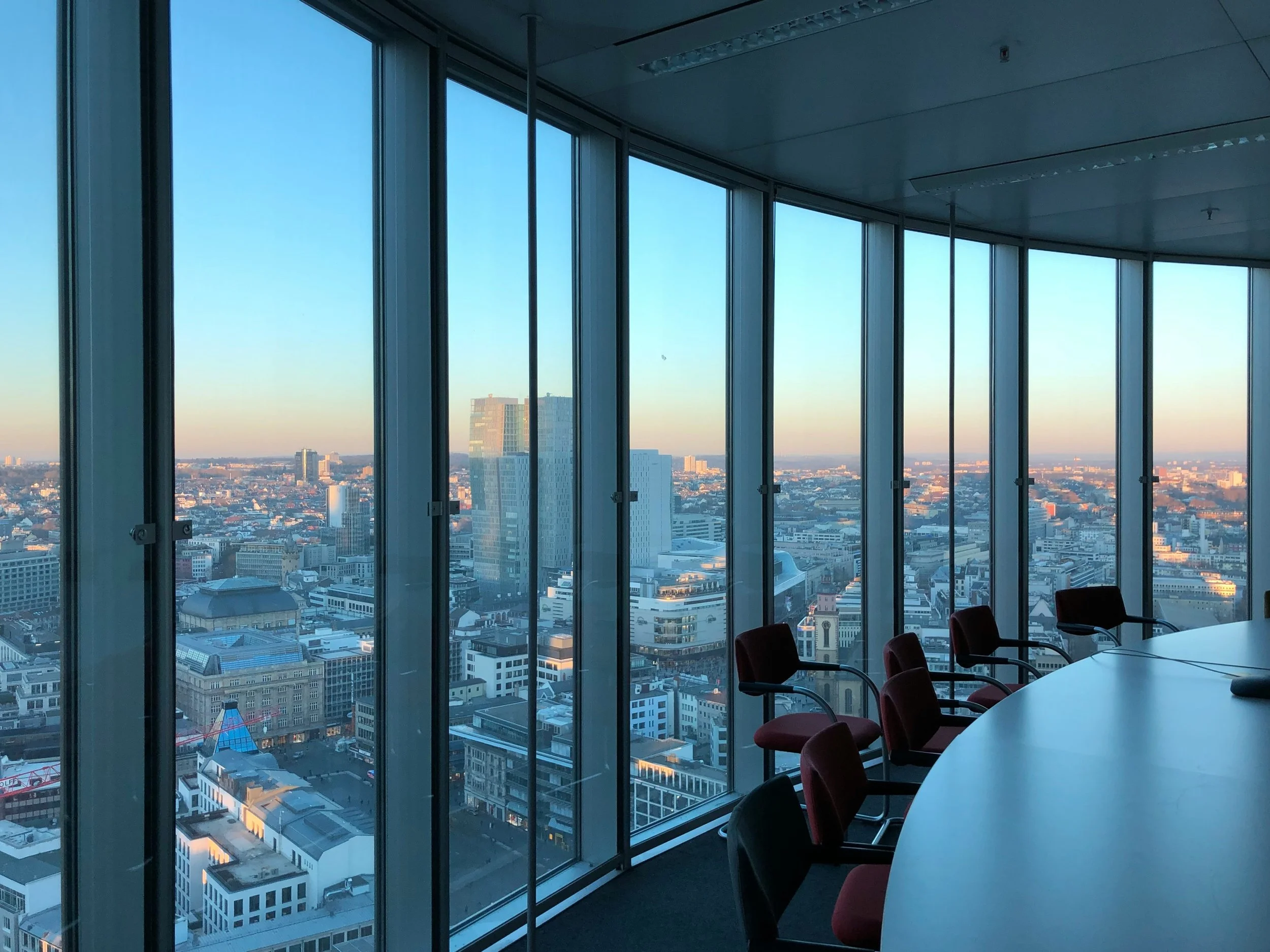 Vista desde una sala de conferencias en un edificio alto, con ventanas de piso a techo que muestran una ciudad moderna con edificios altos y un cielo despejado al atardecer.