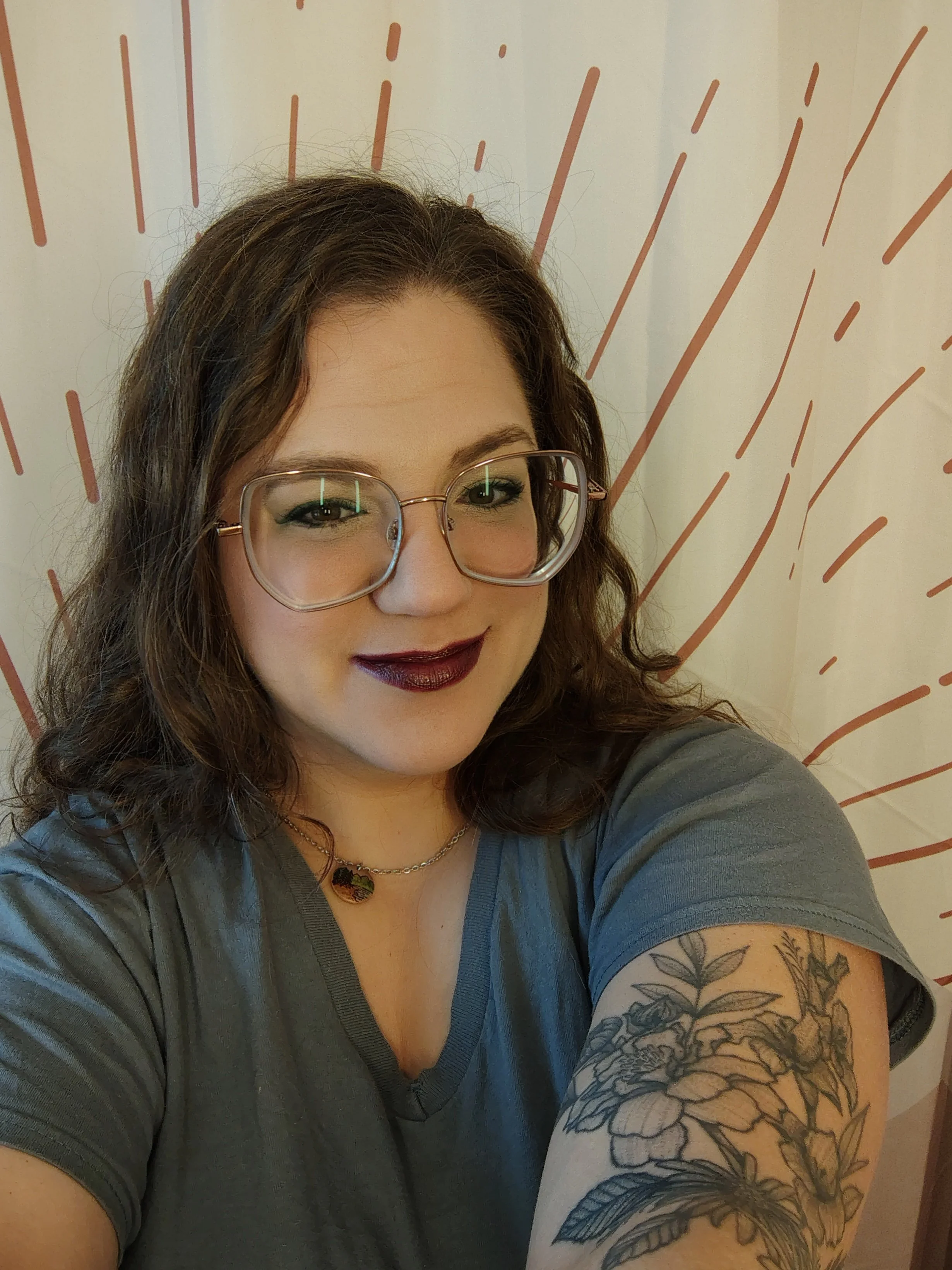 A woman with curly brown hair, light skin, wearing large clear glasses, dark lipstick, a necklace, and a gray T-shirt, smiling in front of a white wall with red abstract lines.