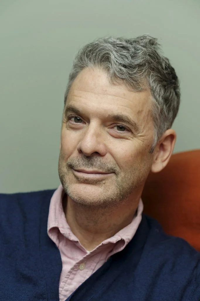 Portrait of a middle-aged man with gray hair, smiling slightly, sitting on a red chair, wearing a pink collared shirt and navy sweater, against a plain background.