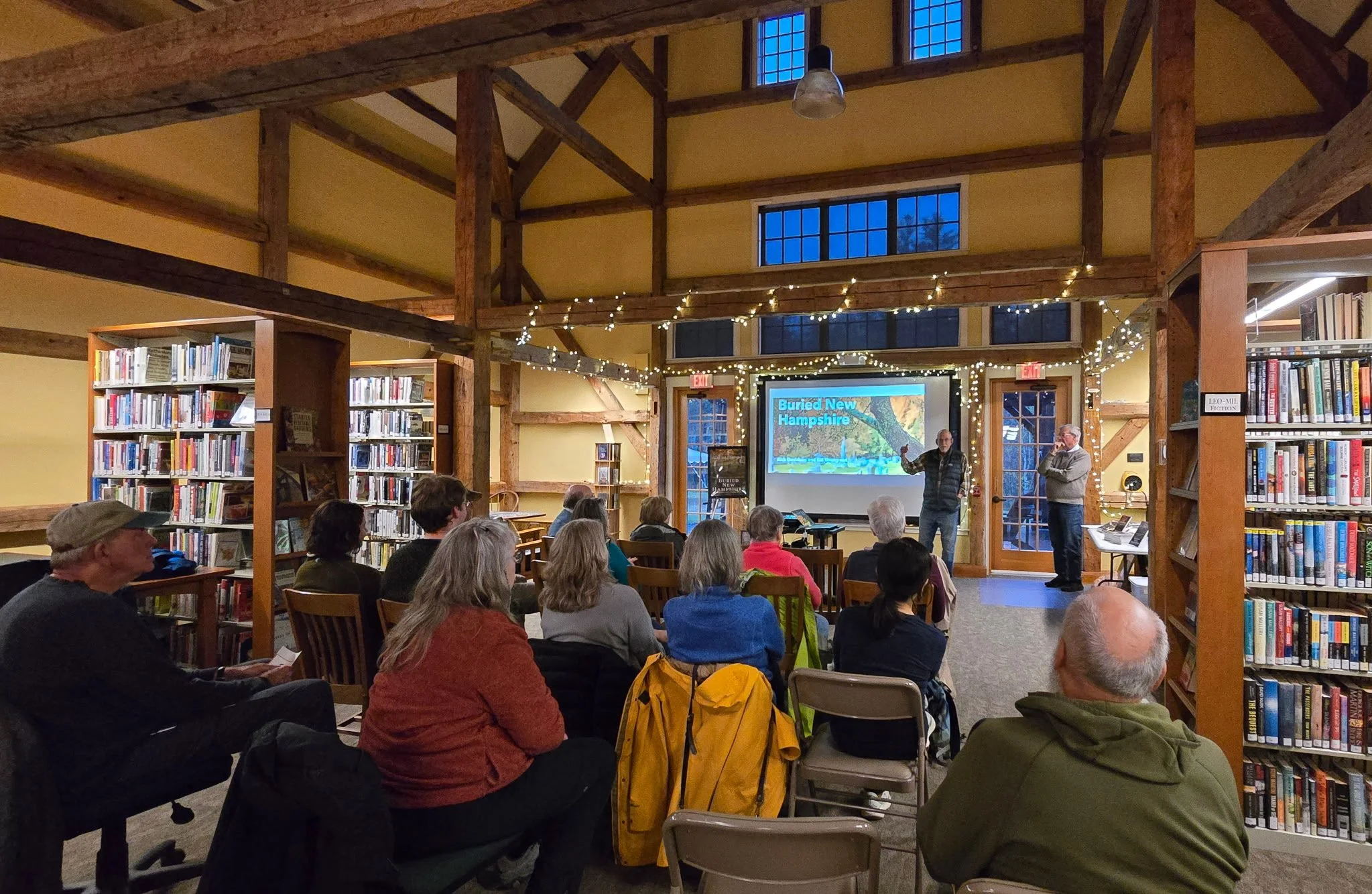 People attending a presentation in a library with wooden beams, bookshelves, string lights, and a large screen displaying the title 'Buried New Hampshire'.