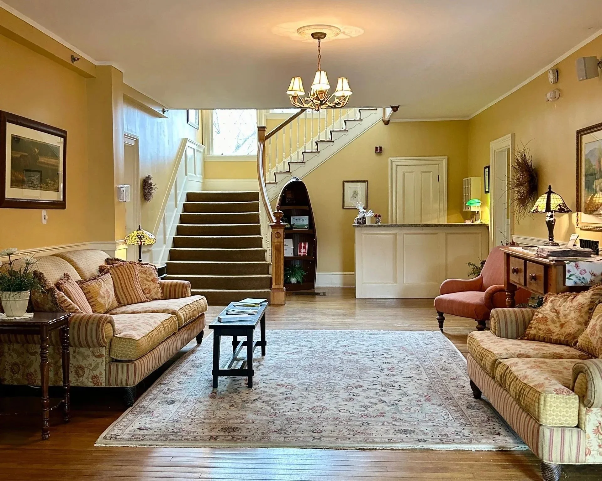 Living room with two patterned sofas, a rug, a coffee table, and a corner with a lamp and decorative items. A staircase with a wooden handrail leads to the upper floor, and a window near the ceiling provides natural light. The walls are painted yellow, and there are framed pictures and decorative wreaths on the walls.