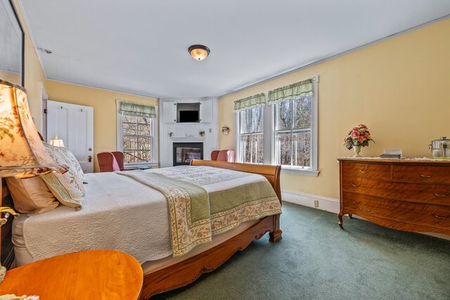 Bedroom with a large wooden bed, beige bedding, a green and beige quilt, and a pillow with floral patterns. There are three windows with curtains, a wooden dresser with a flower vase and books, a small TV above a fireplace, and two red chairs near the corner. The floor is carpeted, and the walls are painted yellow.