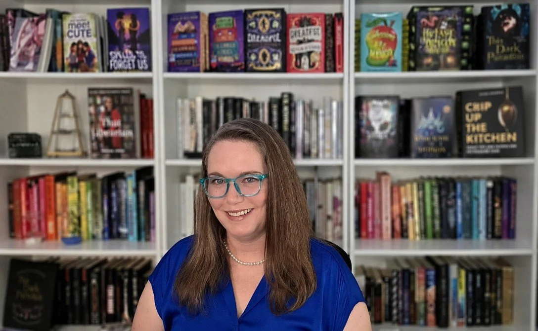A woman wearing blue glasses and a blue top, smiling, sitting in front of a white bookshelf filled with colorful books.