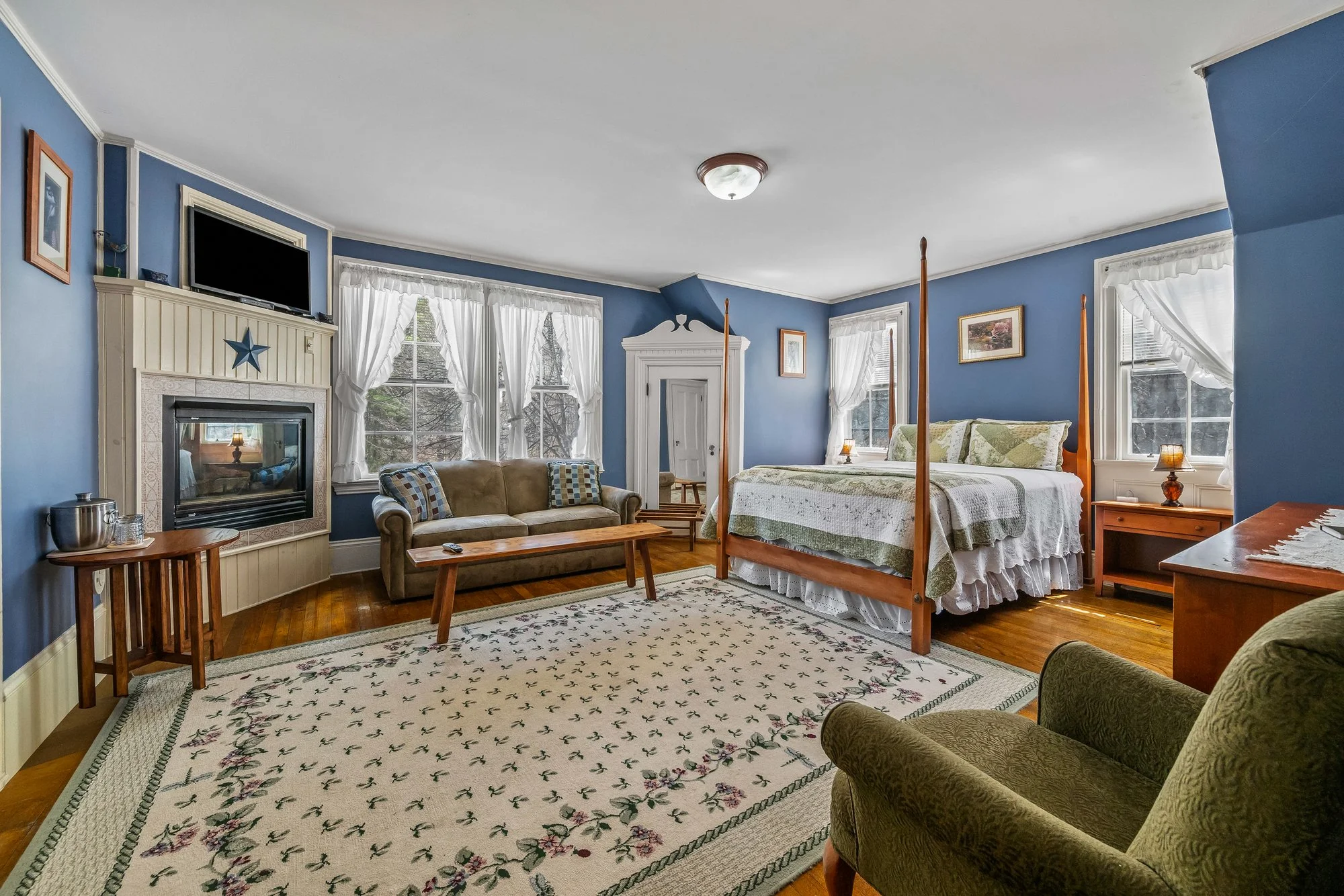 A cozy bedroom with blue walls, white trim, and large windows with white curtains. The room features a four-poster bed with floral bedding, a sofa with patterned pillows, a fireplace with a TV above it, and a wooden coffee table on a white floral rug. There are framed pictures on the walls and a small wooden side table.