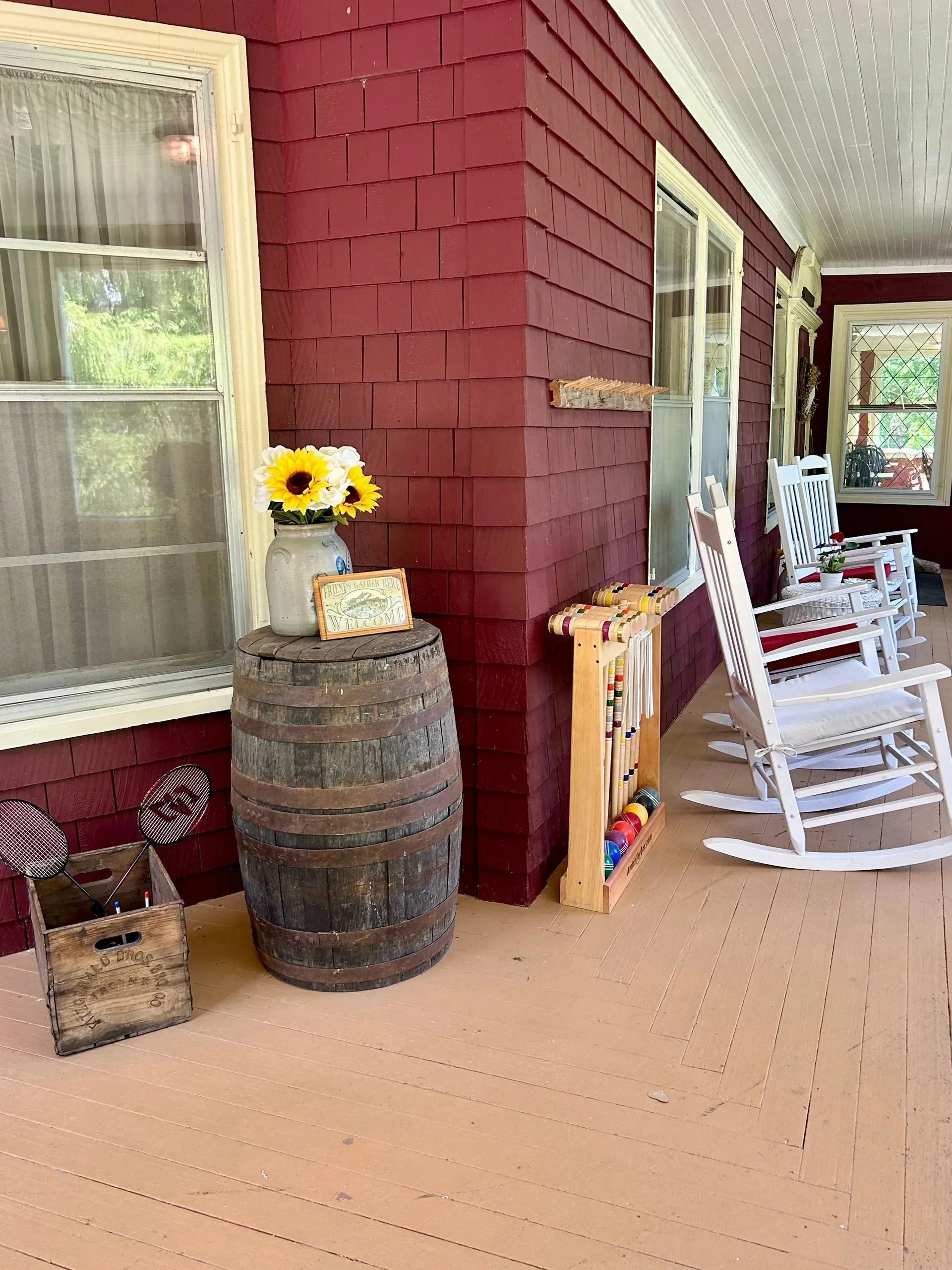 Porch area with red painted wooden exterior, white trim, and a white ceiling. There are four white rocking chairs, a small potted plant, and a wooden golf club rack with colorful golf clubs. A rustic wooden barrel is used as a table, with a vase of sunflowers and a welcome sign on top. A small wooden crate with more golf clubs is on the floor.