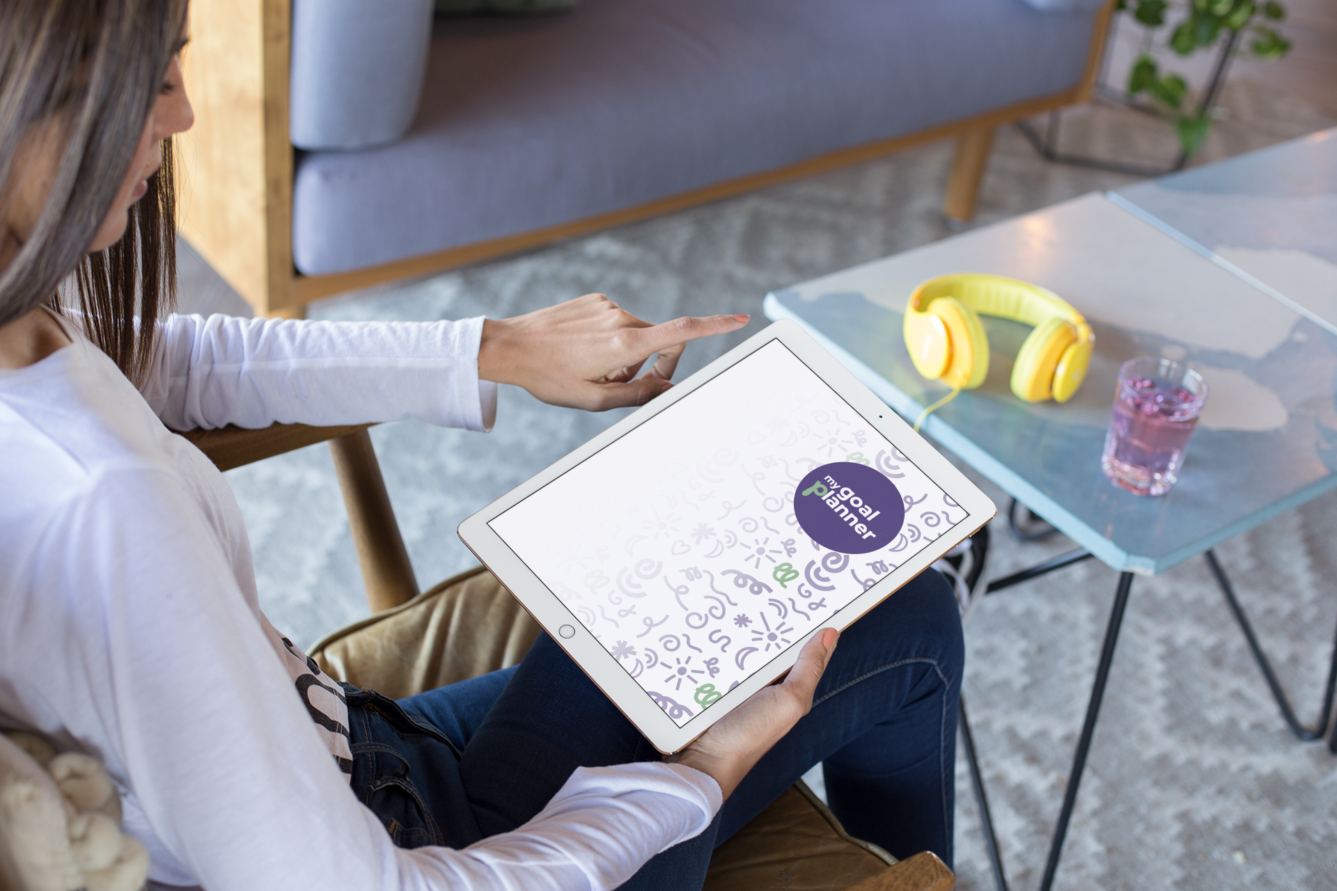 A woman sitting on a couch holding a tablet with a 'goal planner' app on the screen. On the table in front of her are yellow headphones and a glass of pink beverage.