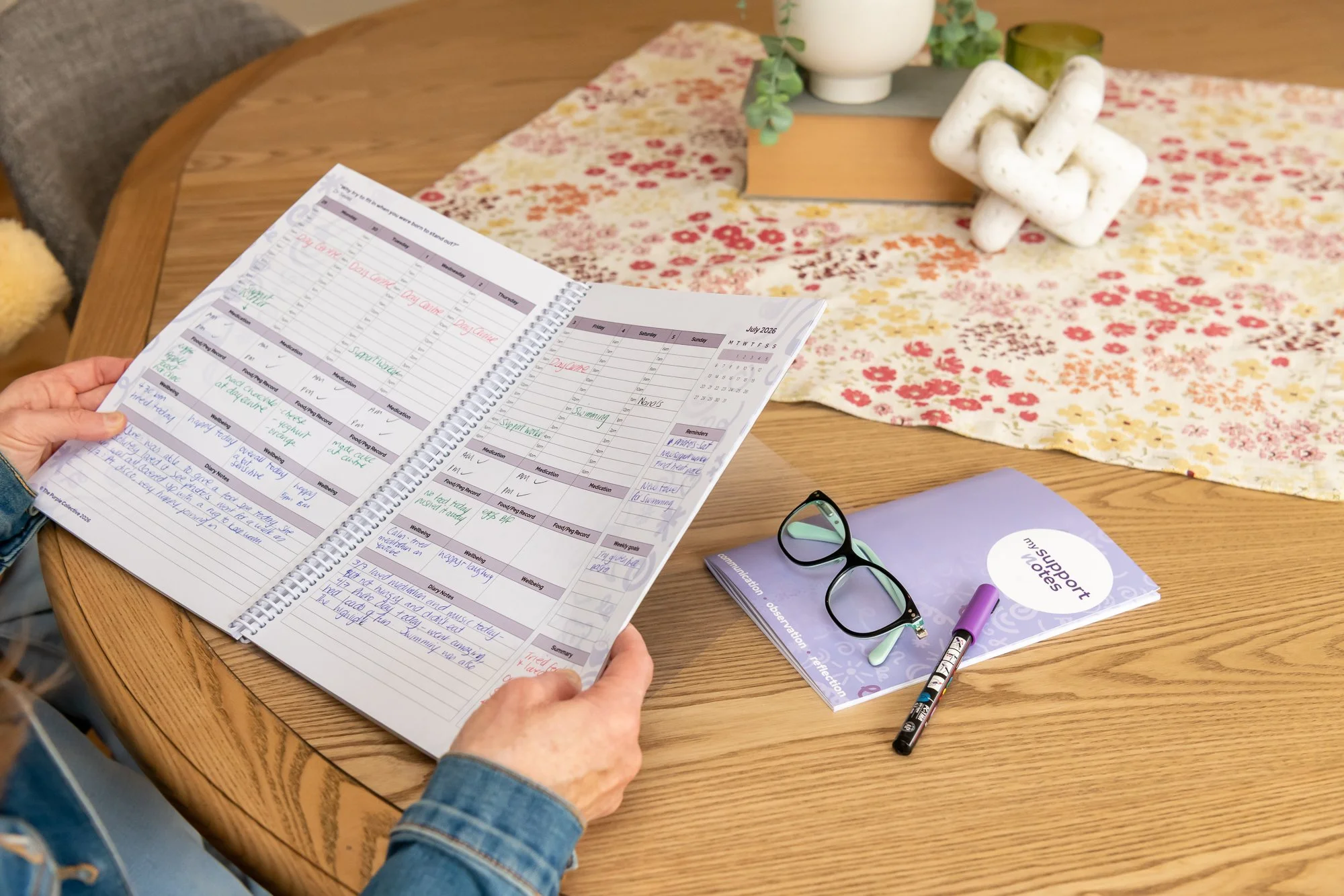 A person's hands holding a weekly planner open on a wooden table. The planner contains handwritten notes and a weekly schedule for July 2026. On the table are a pair of glasses, a purple support notes booklet, a purple marker, and a black marker. The