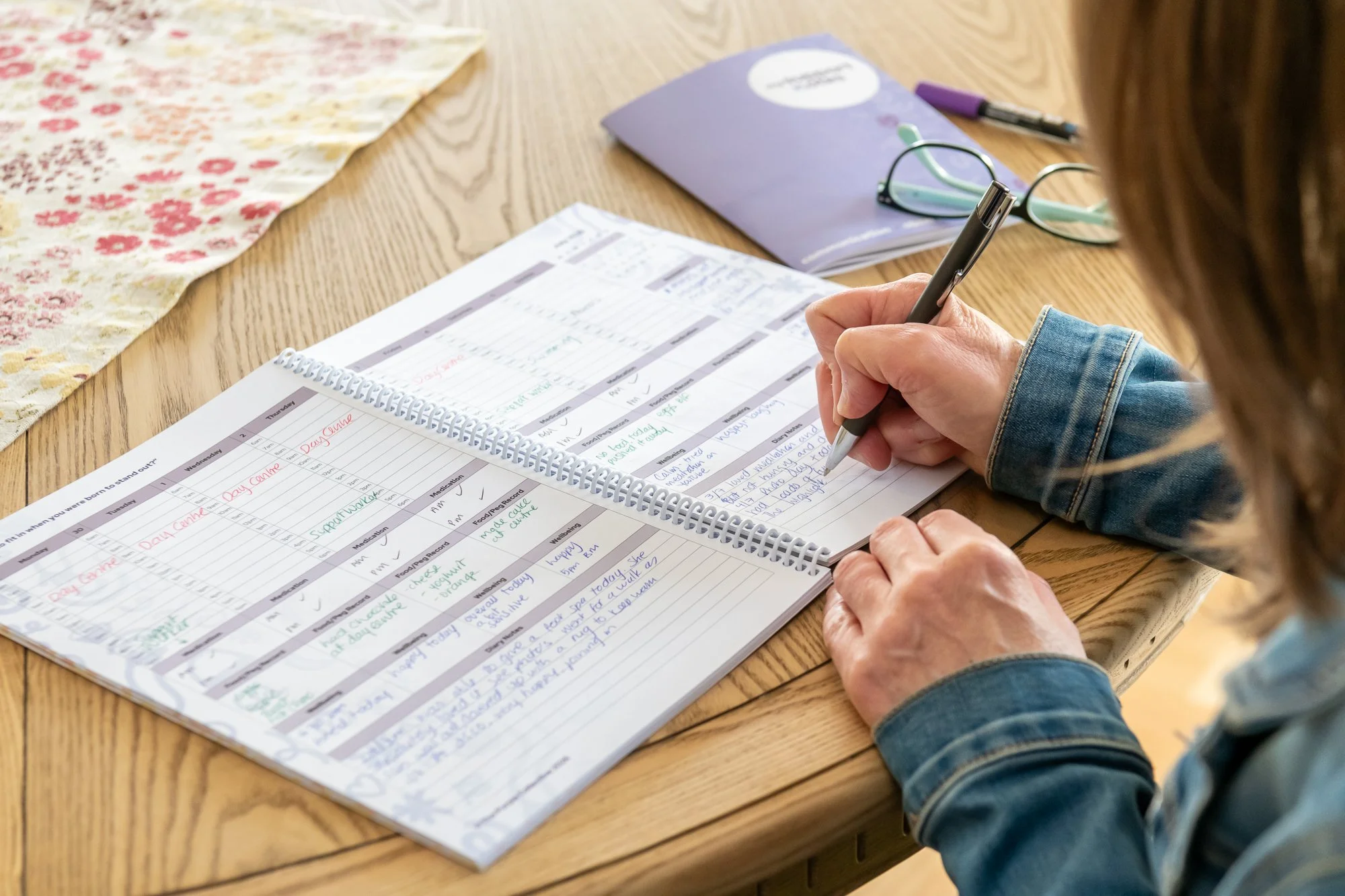 Person wearing a denim jacket writing in a planner with a black pen at a wooden table, with glasses, a purple folder, a pen, and a floral fabric on the table.