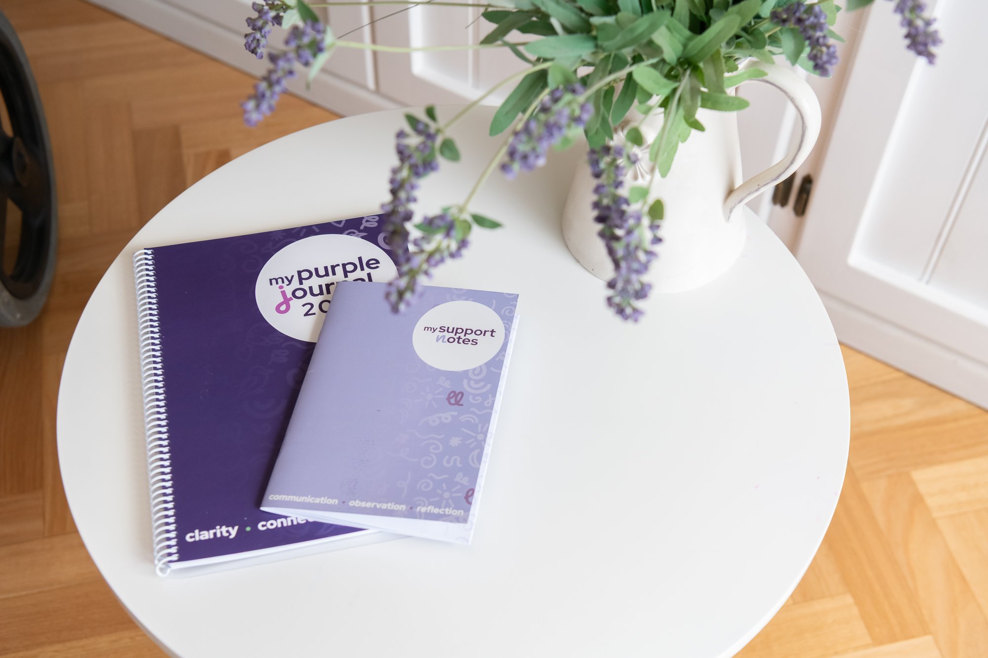 A white round table with purple and white notebooks titled 'my purple journal 2023' and 'my support notes,' placed next to a white pitcher filled with purple lavender flowers, in front of a white door.