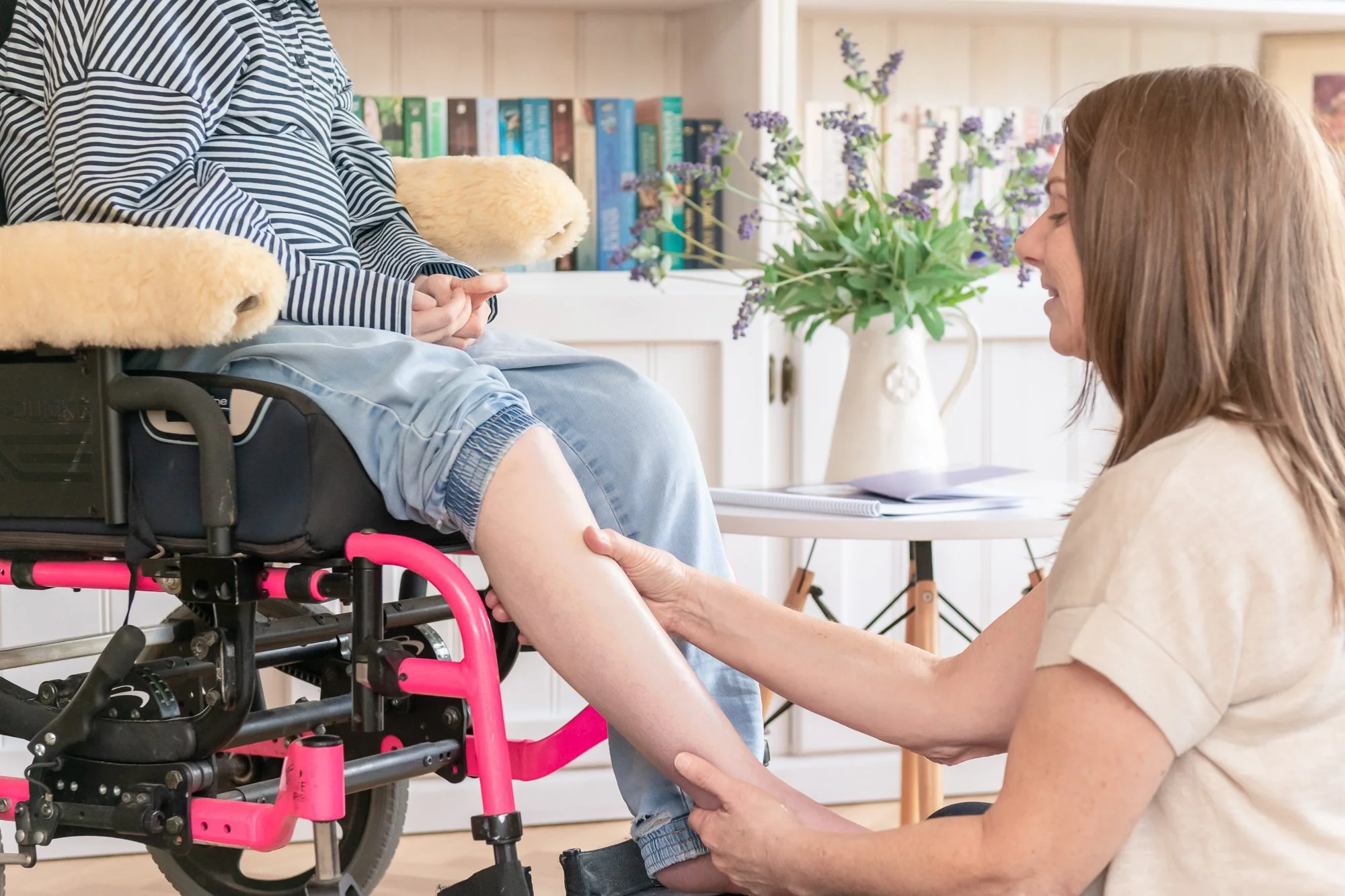 A woman helping a person in a wheelchair by holding their foot. The background shows a bookshelf, a table with purple flowers, and a cozy, bright room.