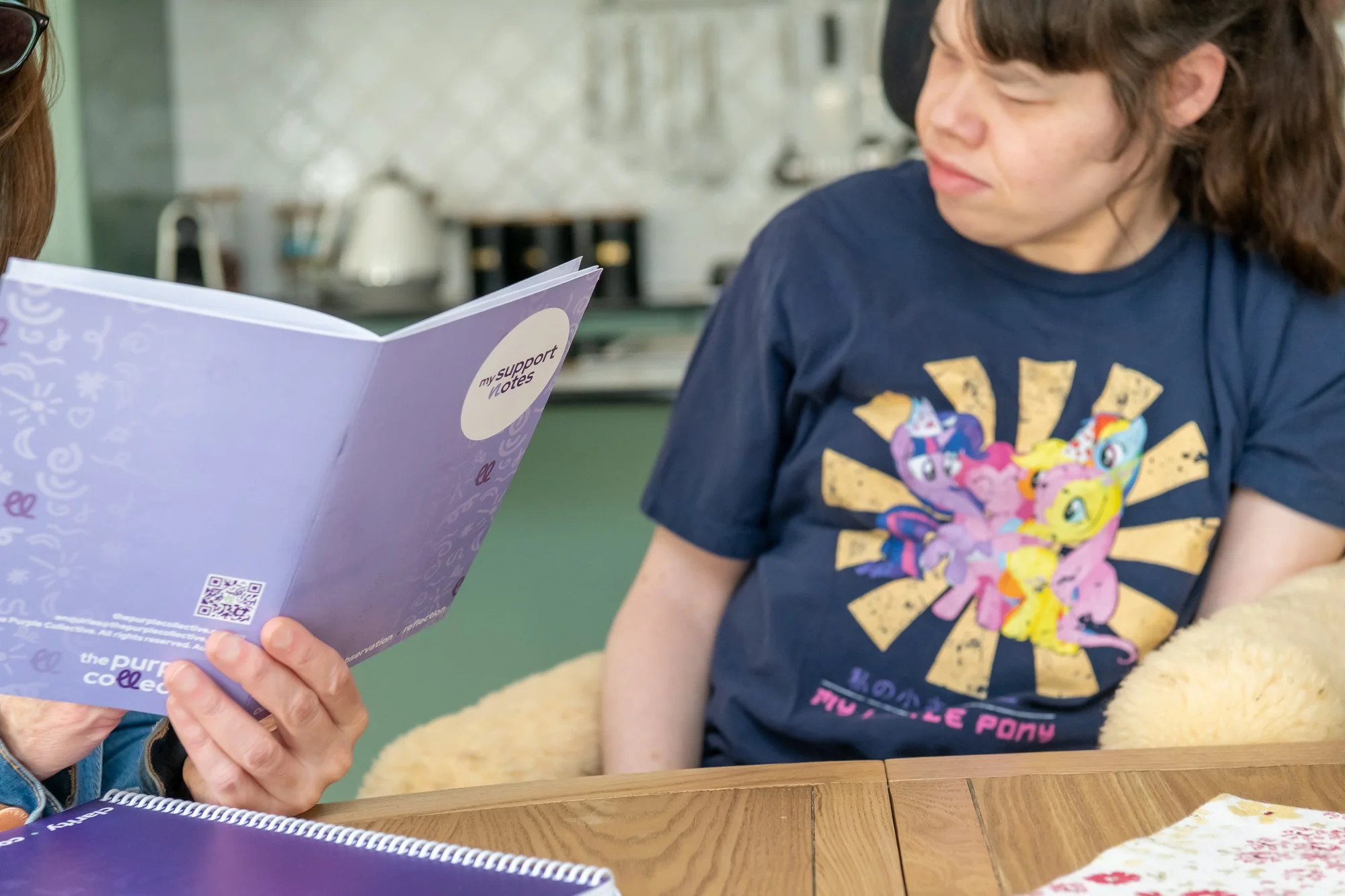 A person holding a lavender notebook titled 'my support notes' with a QR code on the bottom, while another person wearing a My Little Pony T-shirt looks on in a casual indoor setting.