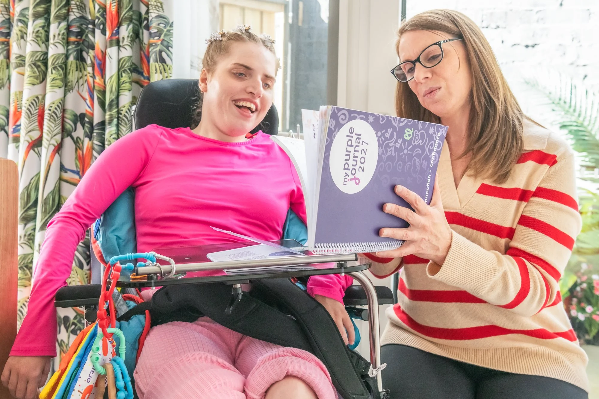 A young girl in a wheelchair with pink clothing smiling and talking to an older woman with glasses and a striped sweater, who is reading a book together in a bright room with floral curtains and large windows.