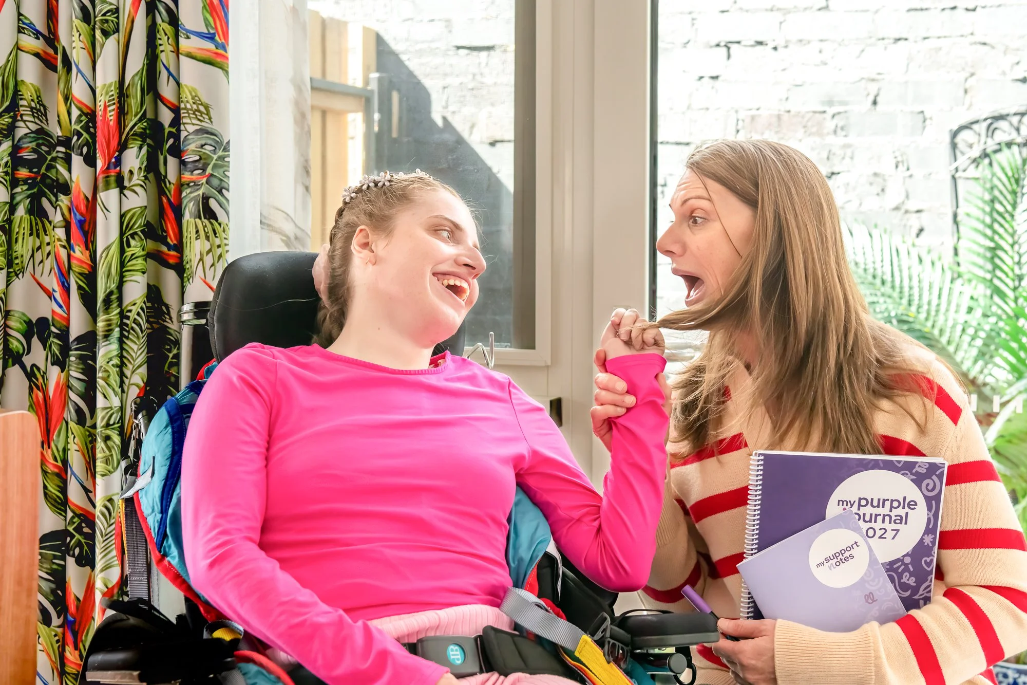 A young girl in a pink long-sleeve shirt in a motorized wheelchair, smiling and engaging with an older woman who is holding her hand and speaking to her. The woman has long brown hair and is wearing a red and white striped sweater. They are indoors near a window with a decorative colorful curtain on the left, and a white brick wall in the background.