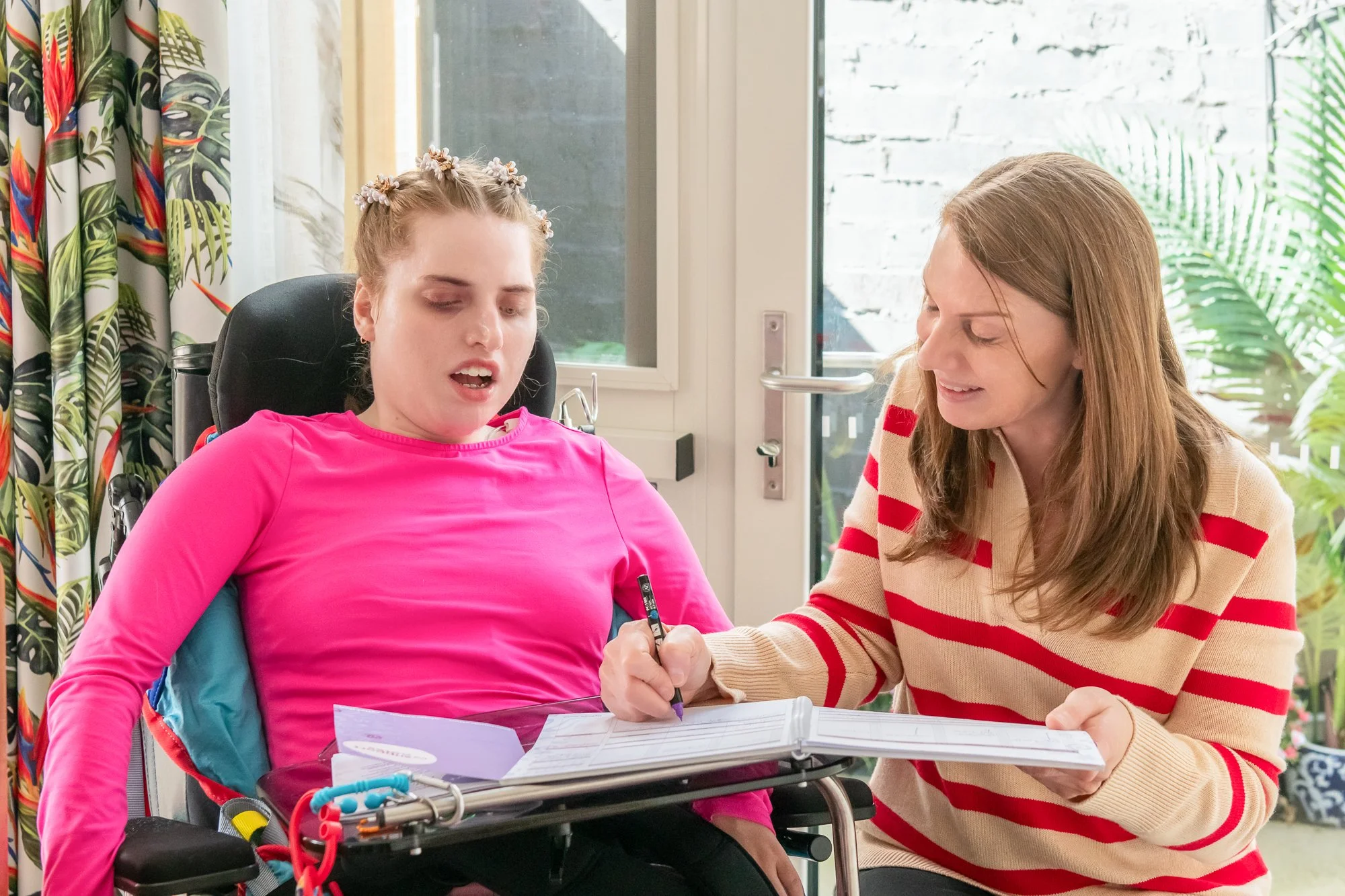 A woman with long brown hair and a striped red and cream sweater is sitting at a table, writing in a notebook. A young woman with light hair styled with small flowers, wearing a bright pink shirt, is sitting in a wheelchair nearby. The wheelchair has