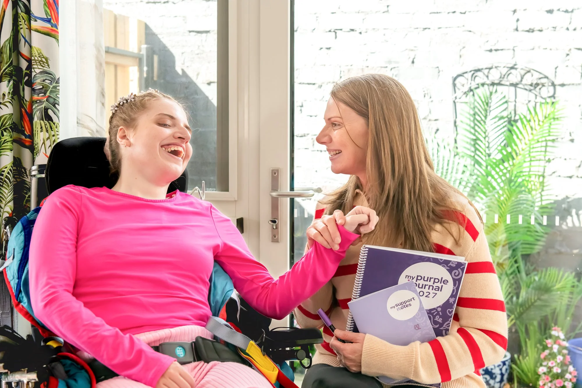 A girl in a pink long-sleeve shirt and a woman smiling, holding hands in a supportive gesture, sitting near a window with white brick wall and green plants outside. The girl is in a wheelchair, and the woman is holding a notebook with