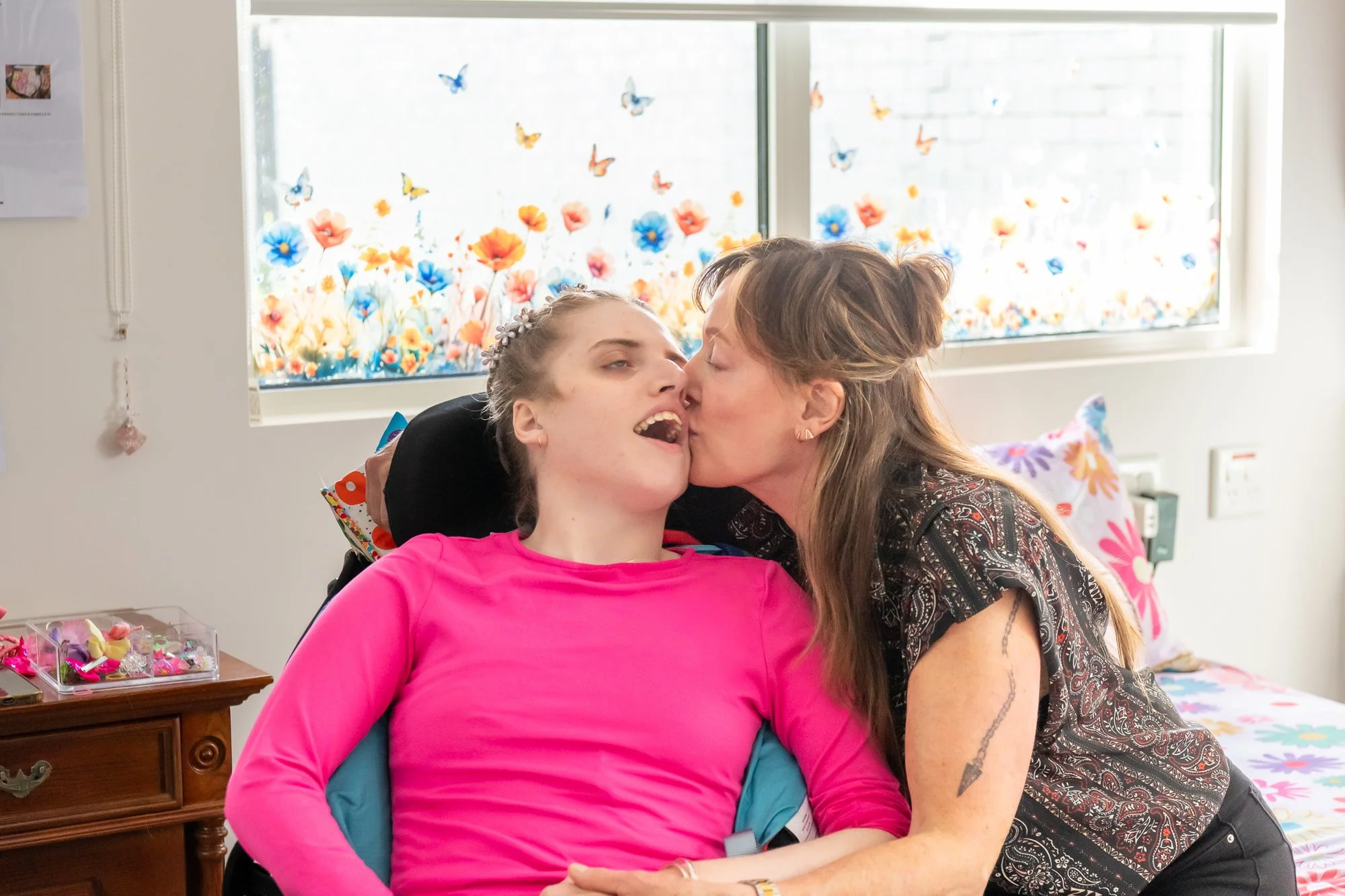 A young woman with a floral headband and a pink shirt is sitting in a wheelchair. An older woman is leaning in to kiss her on the lips. They are in a room decorated with colorful paper butterflies and flowers on a window.