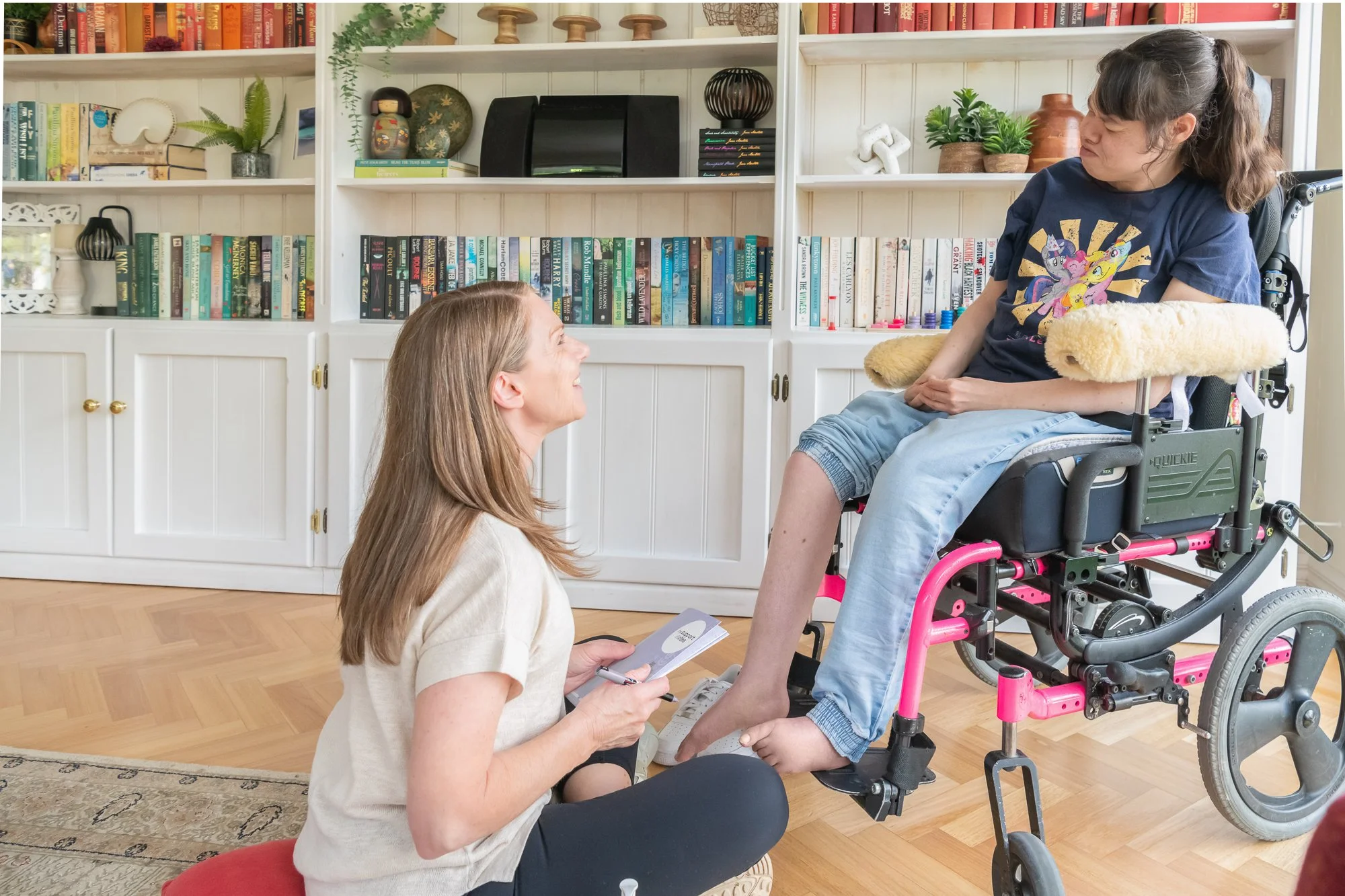 A woman with long hair kneels on the floor in front of a young girl in a wheelchair, holding a book or tablet. The girl has curly hair and is looking at the woman with a playful expression. They are in a room with white bookshelf filled with books an