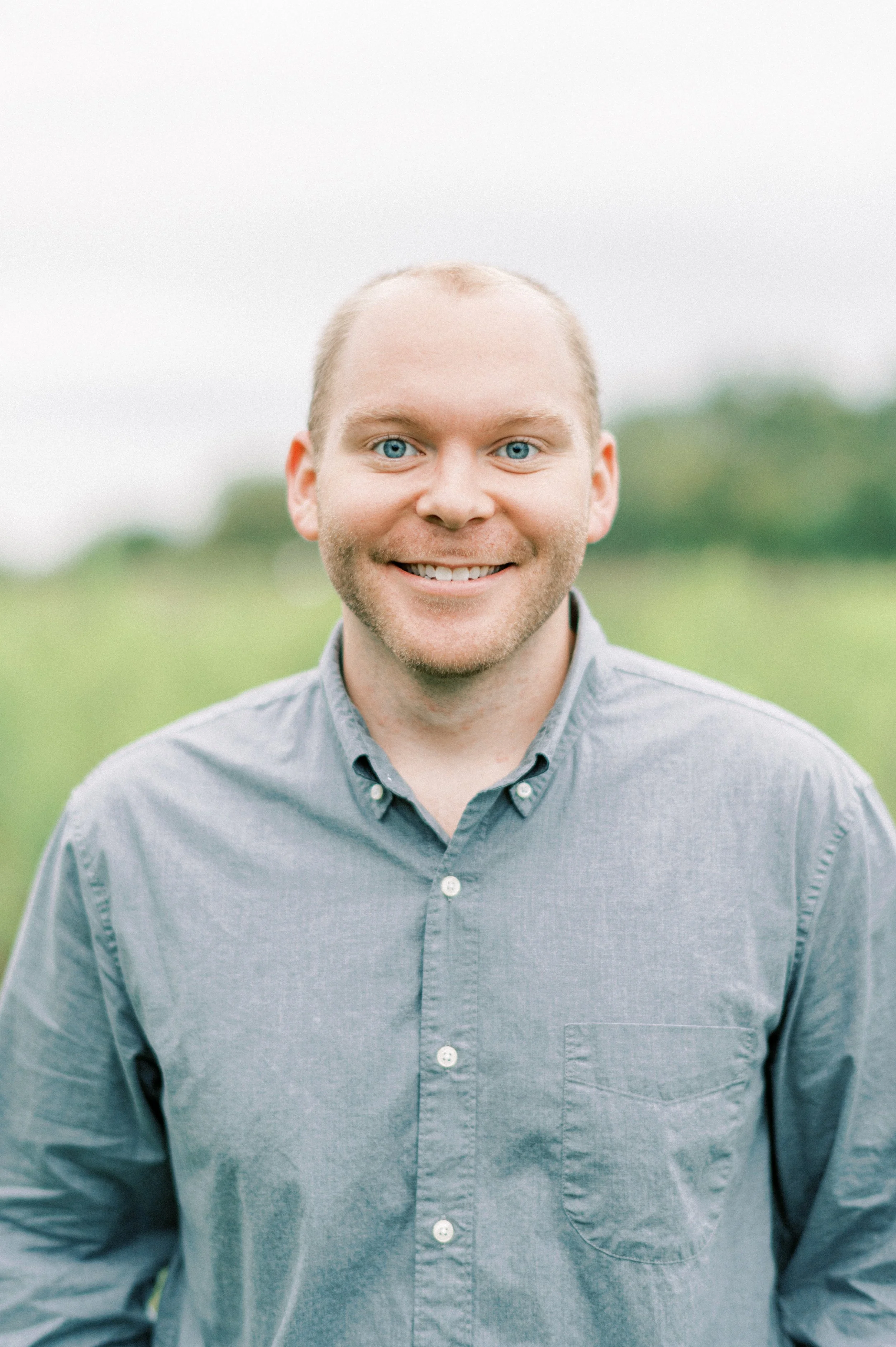 Smiling man with blue eyes and short hair, wearing a gray button-up shirt, standing outdoors in a green field with a cloudy sky.