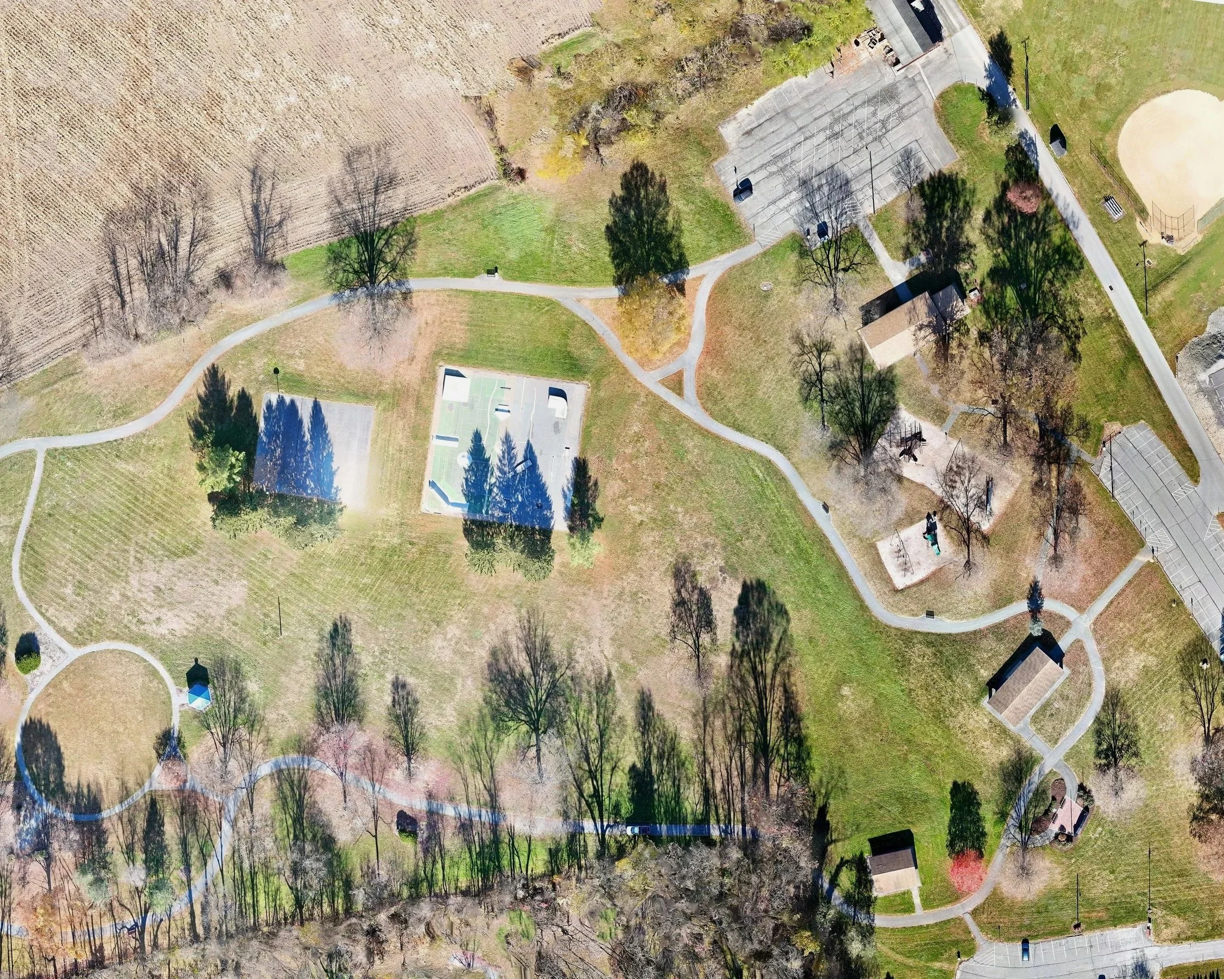 Aerial view of a park with walking paths, tennis courts, open grassy areas, trees, small structures, and parking lots.