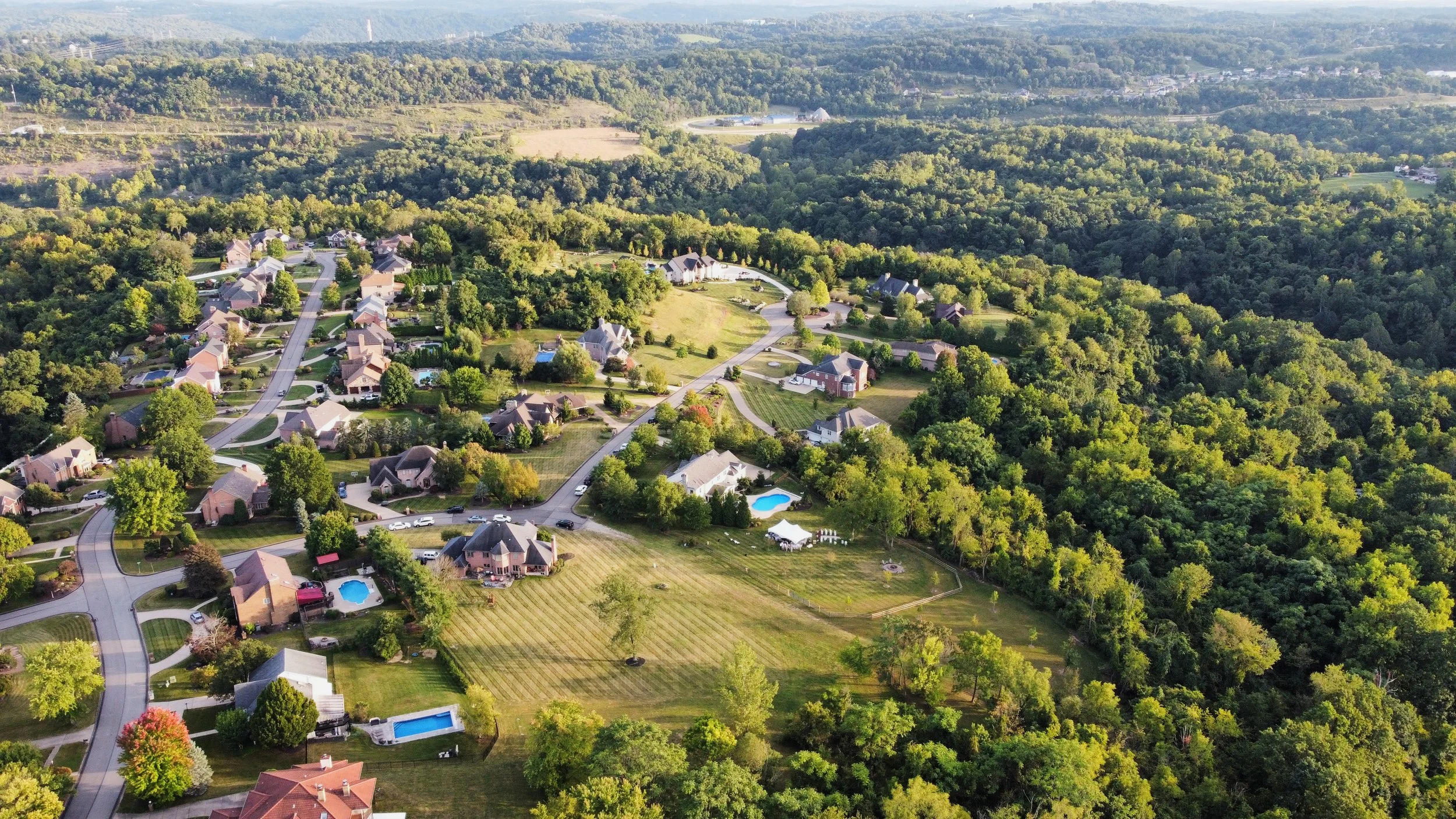 Aerial view of a suburban neighborhood with houses, trees, and swimming pools, surrounded by lush green forest.