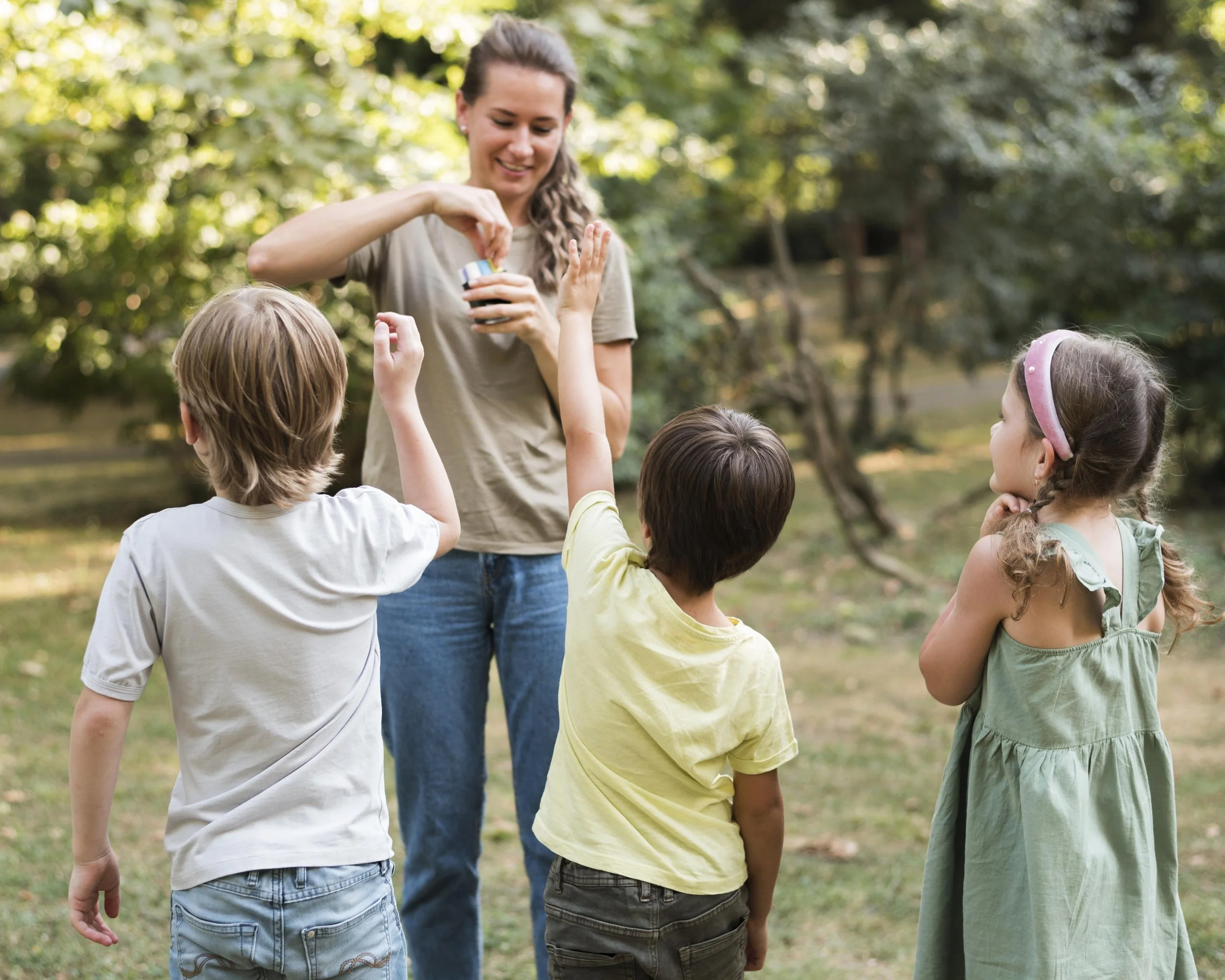 A babysitter and four children in a park, with the woman giving a treat to one of the children.