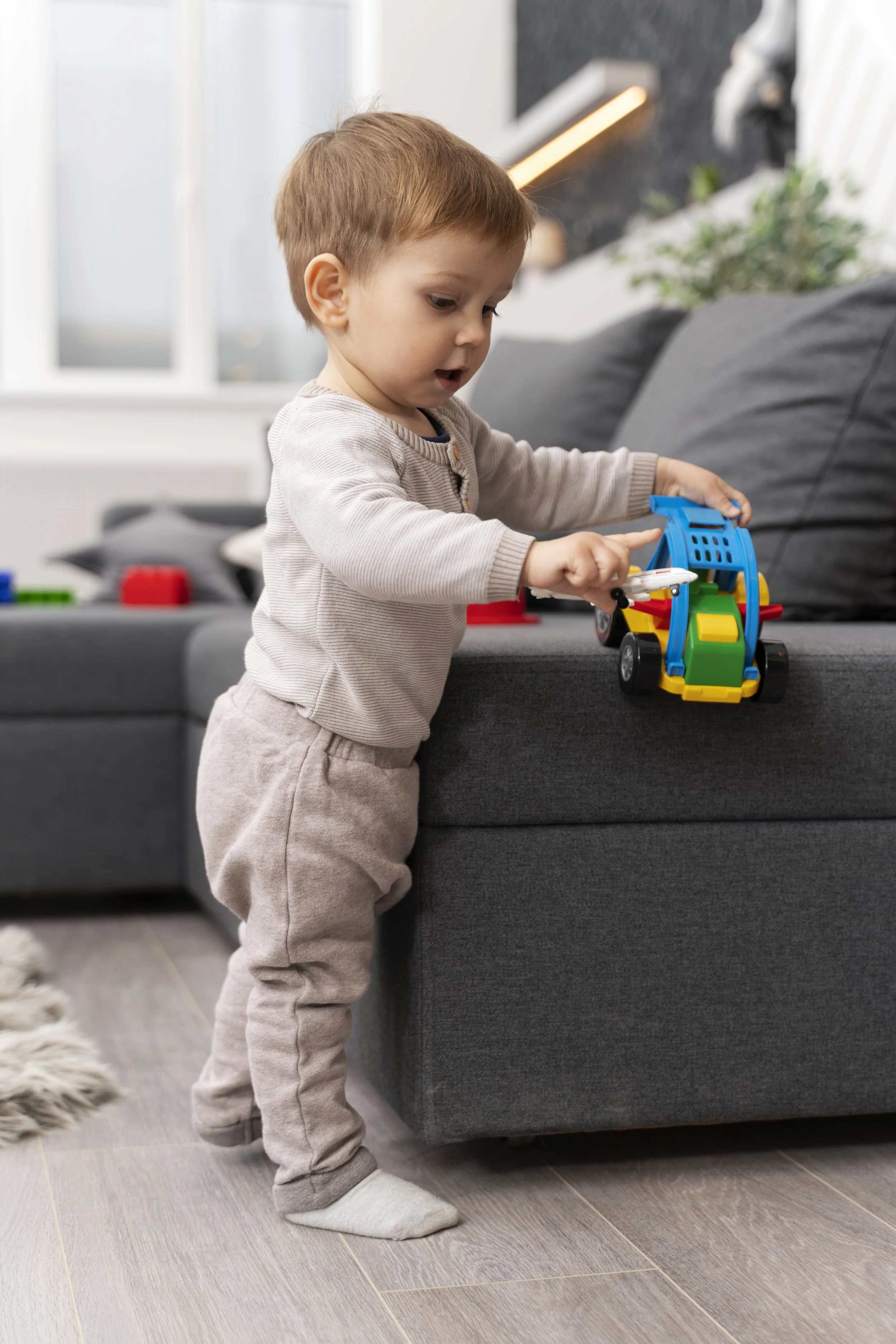 Little boy standing by a couch playing with toy truck.